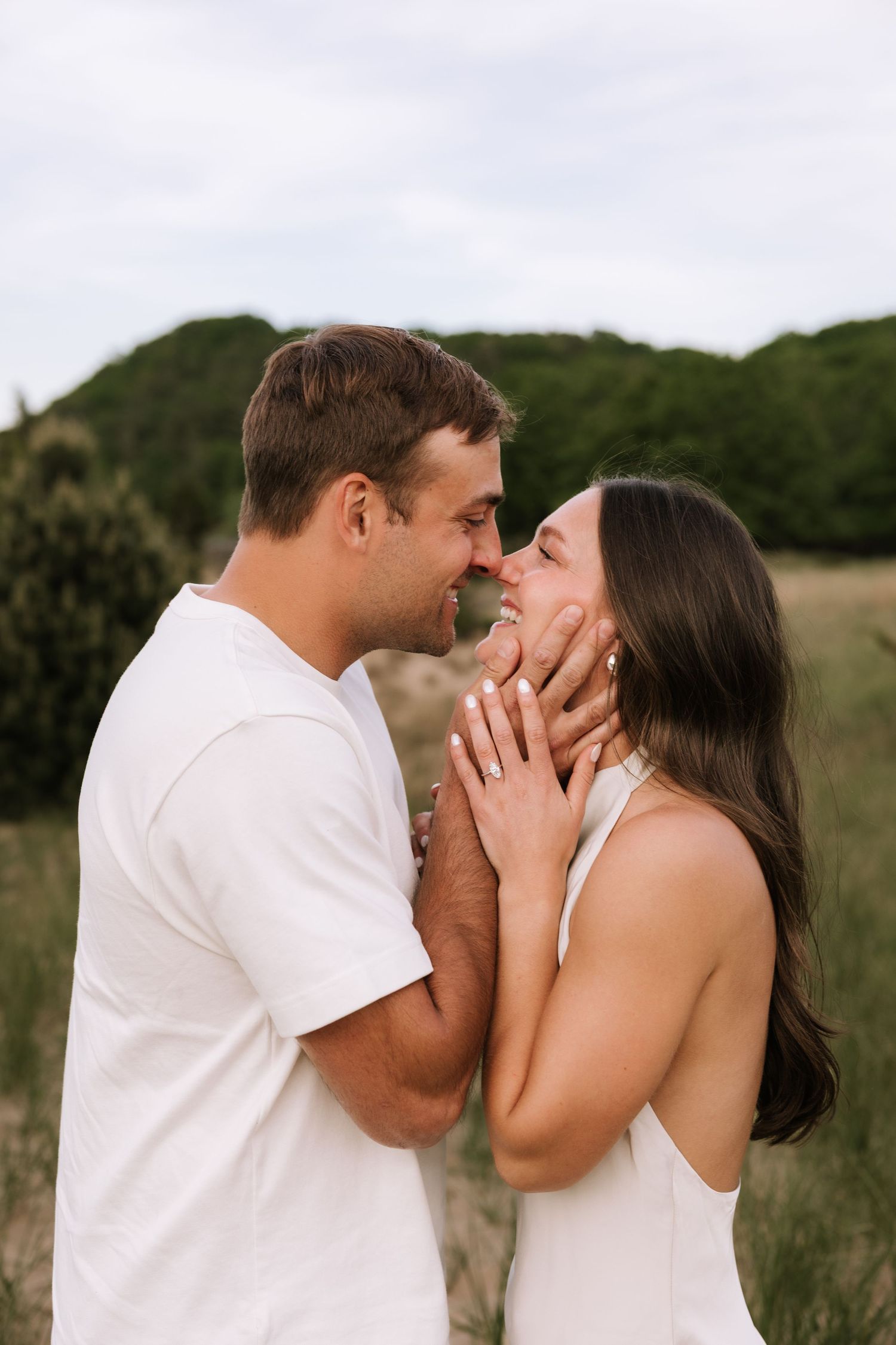 Couple in white outfits sharing a romantic moment in a field, man gently holding woman's face as they lean in to kiss.