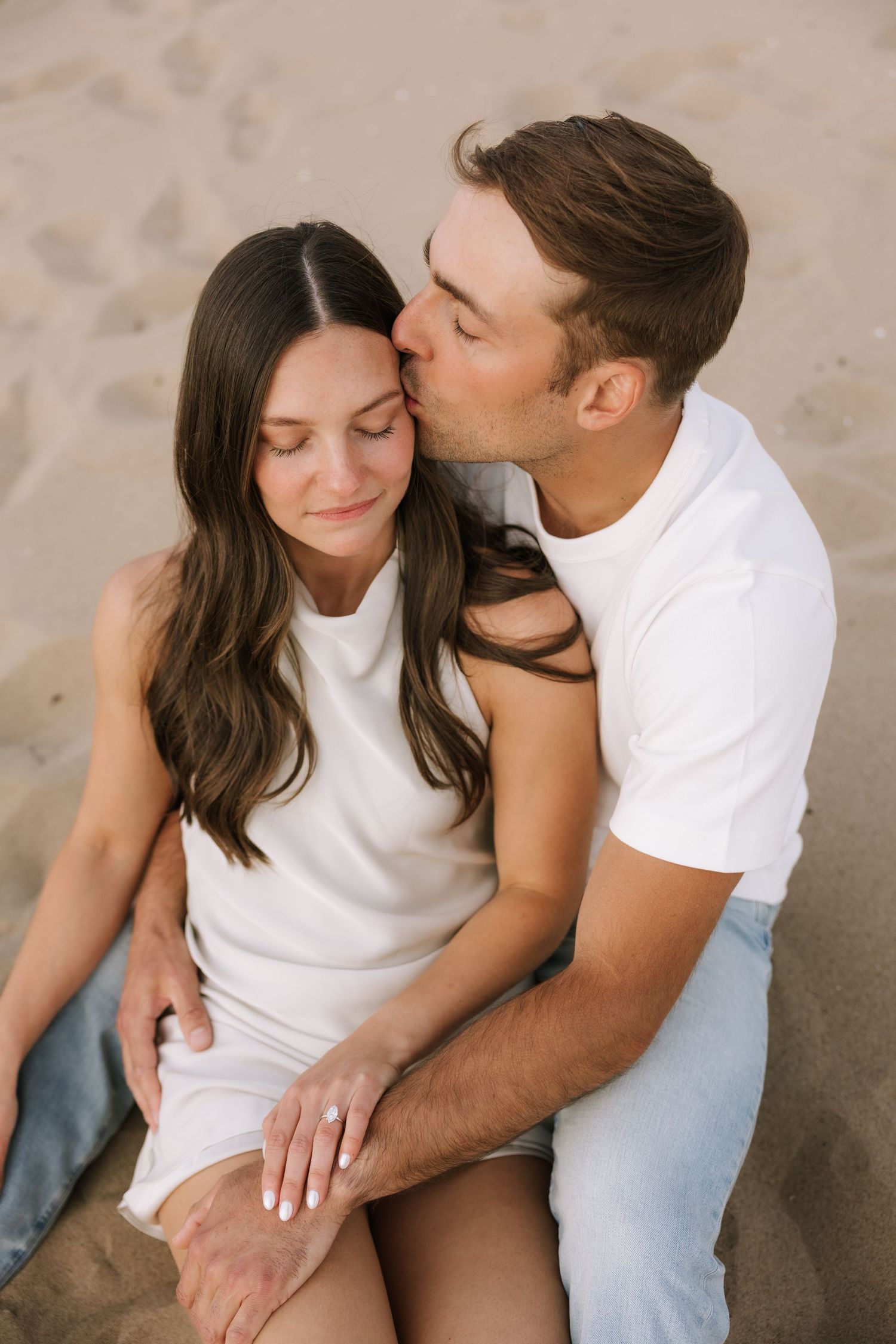 Man kissing woman's cheek while sitting on beach sand, woman showing engagement ring, both in white outfits.