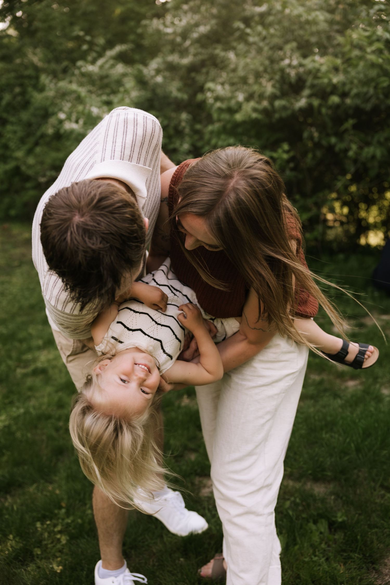Parents playfully dipping their laughing blonde child upside down in a lush green backyard.