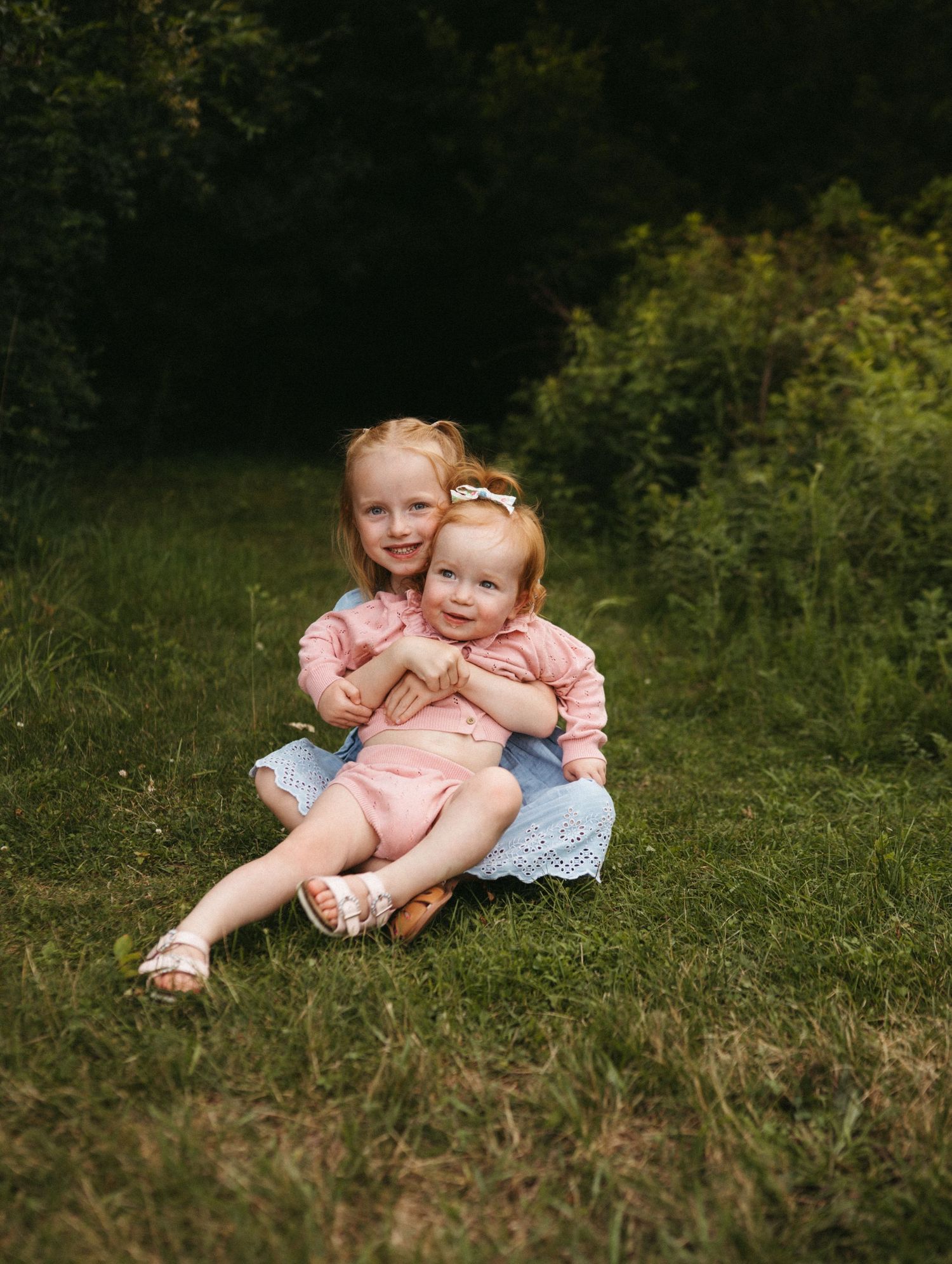 Two young children in matching pink outfits sit together on grass during an outdoor photo session.