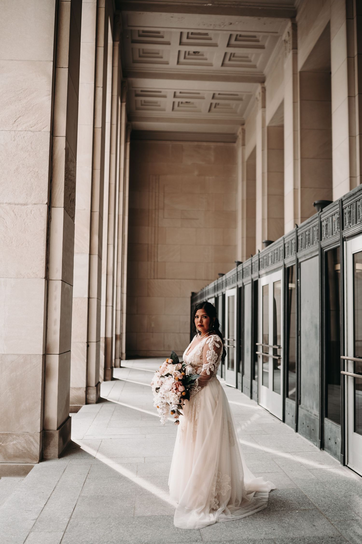 A bride in a lace wedding gown poses elegantly in a columned hallway with a cascading bouquet.
