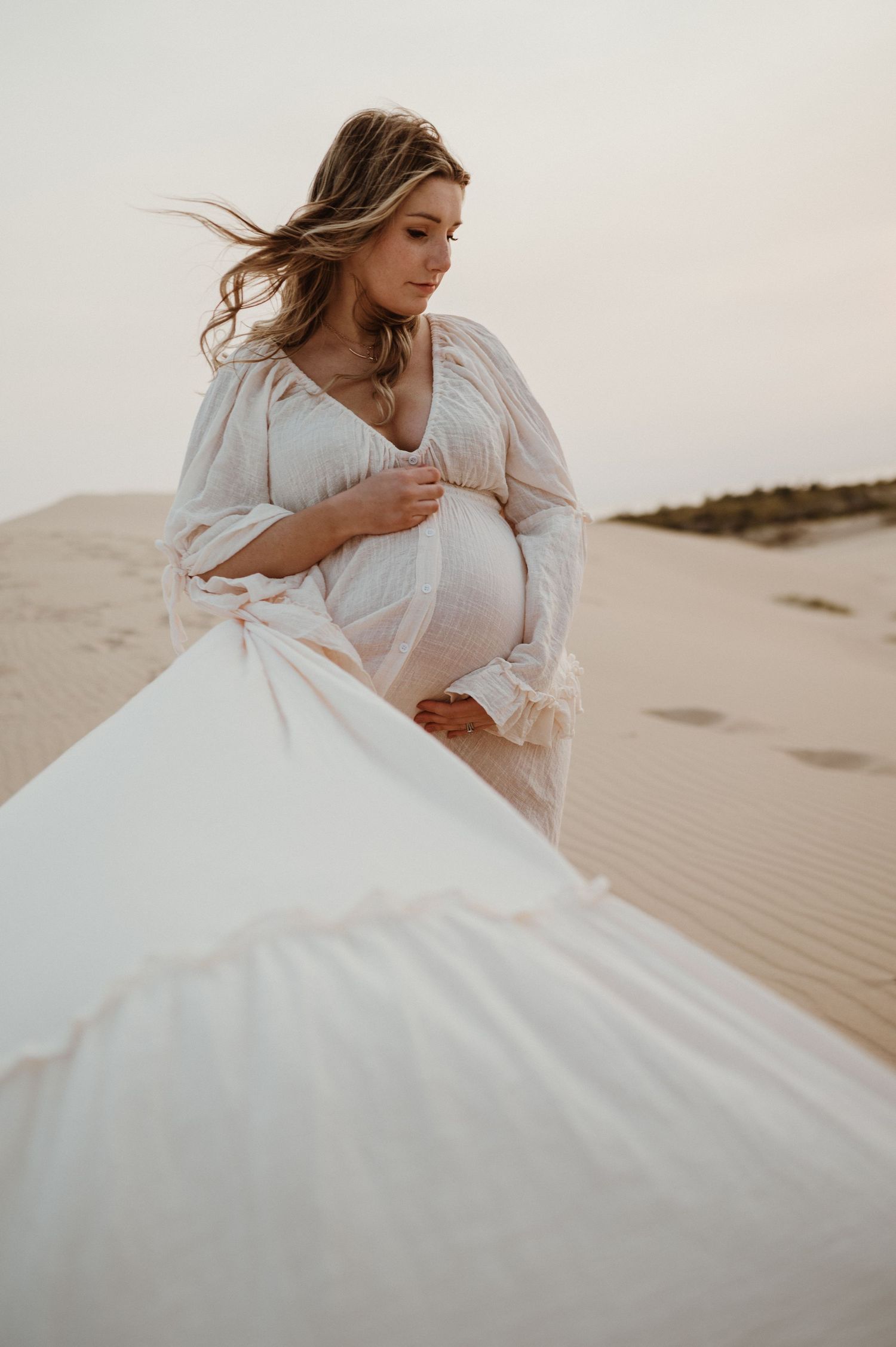 An expectant mother in white dress cradles her belly while standing on desert sand dunes.