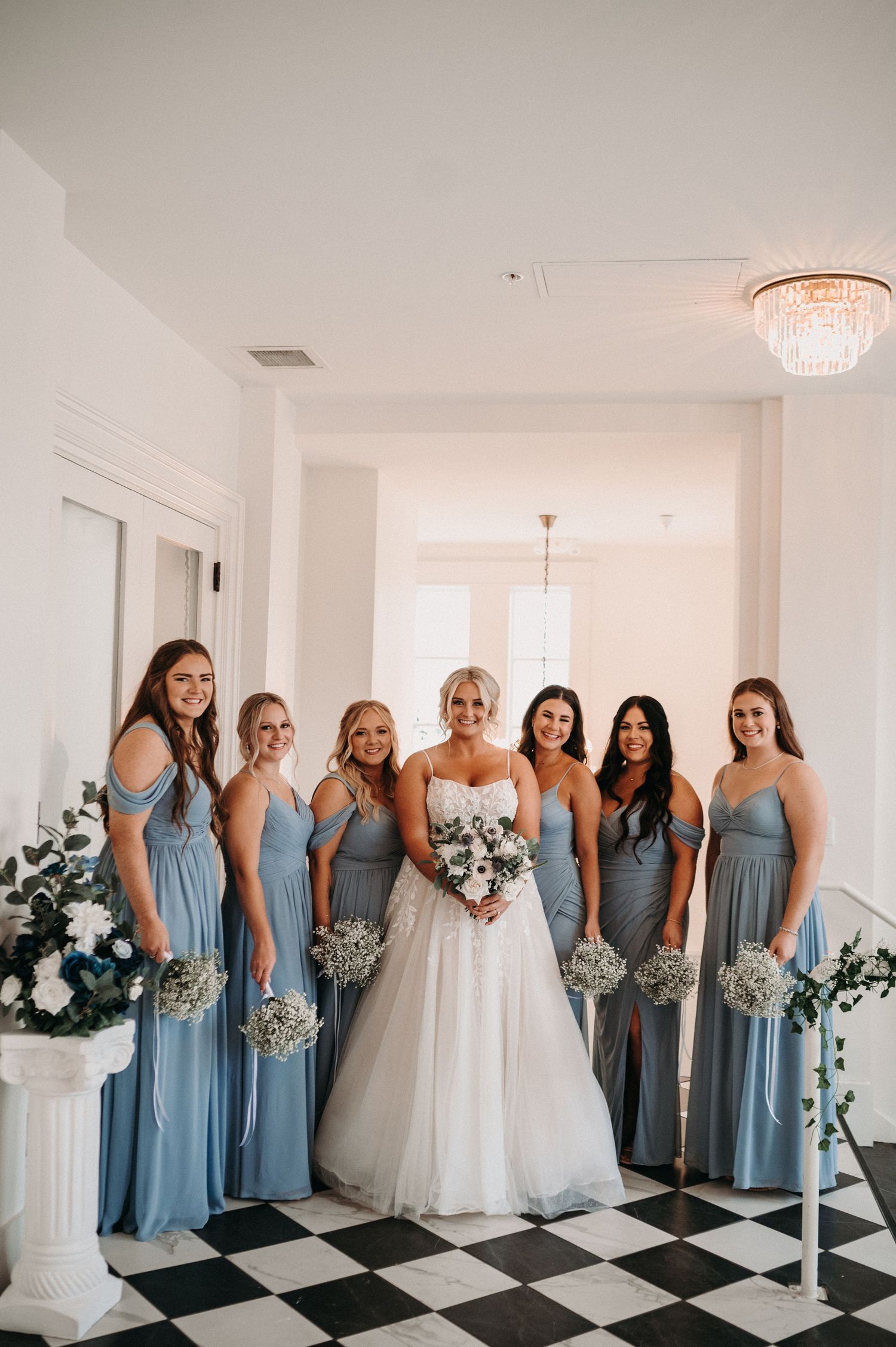 Bridal party in dusty blue dresses poses on black and white checkered floor under crystal chandelier.