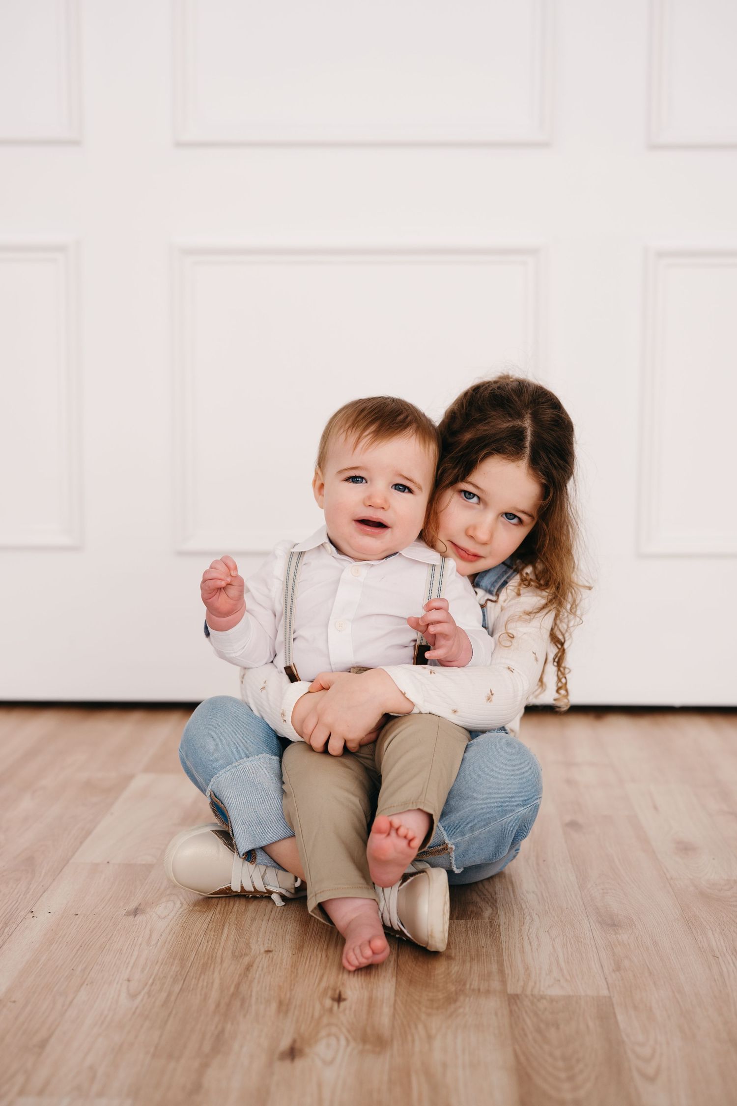 Sweet sibling moment captured on hardwood floor against white paneled wall.