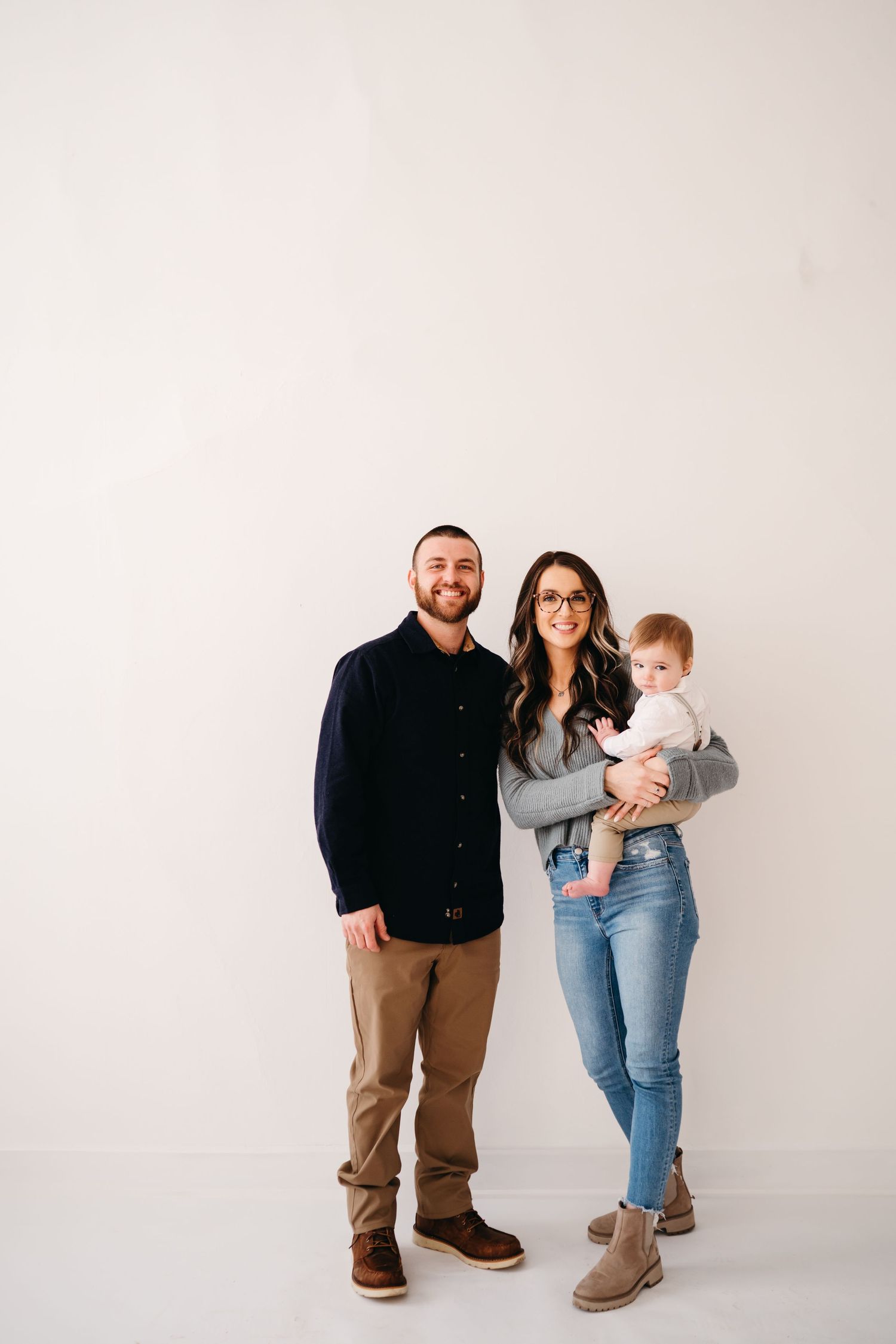 A family stands together against a white backdrop wearing casual clothing while holding a young child.