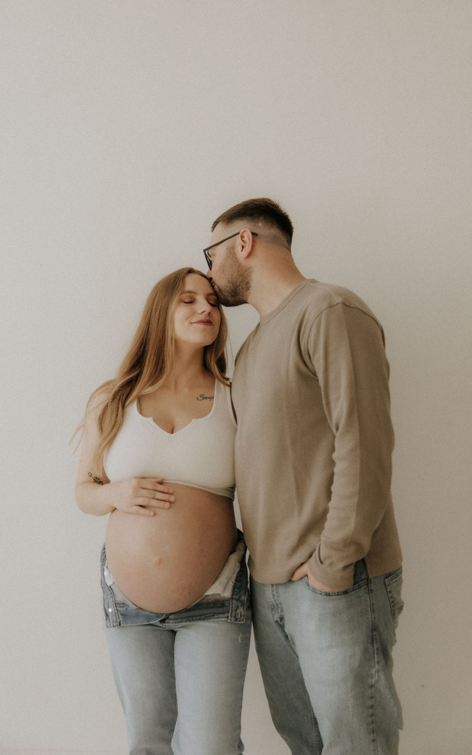 Artistic maternity portraits of couple in matching denim and neutral tones against white wall.