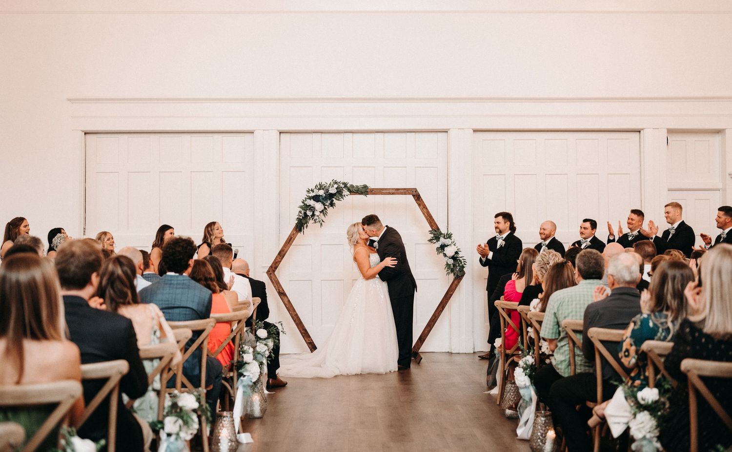 Indoor wedding ceremony with geometric arch and floral decorations as guests watch from wooden chairs.