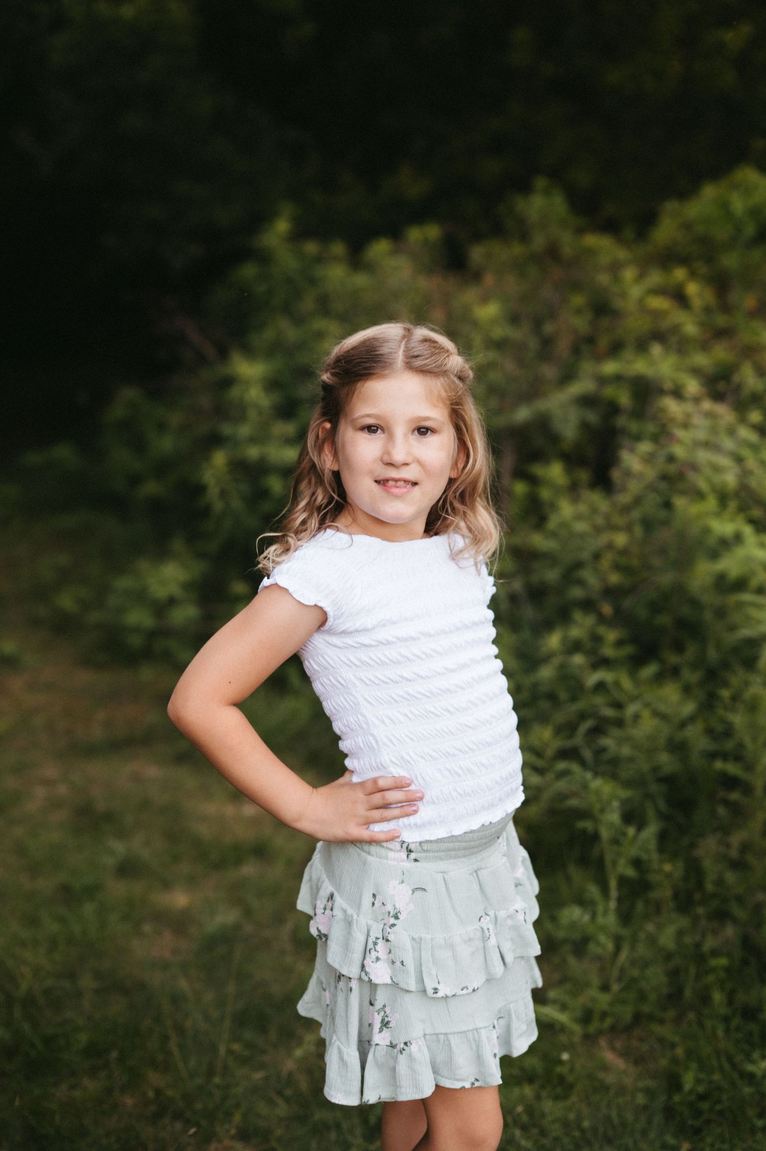 Child poses confidently in white ruffled top and mint green skirt during outdoor photo session.