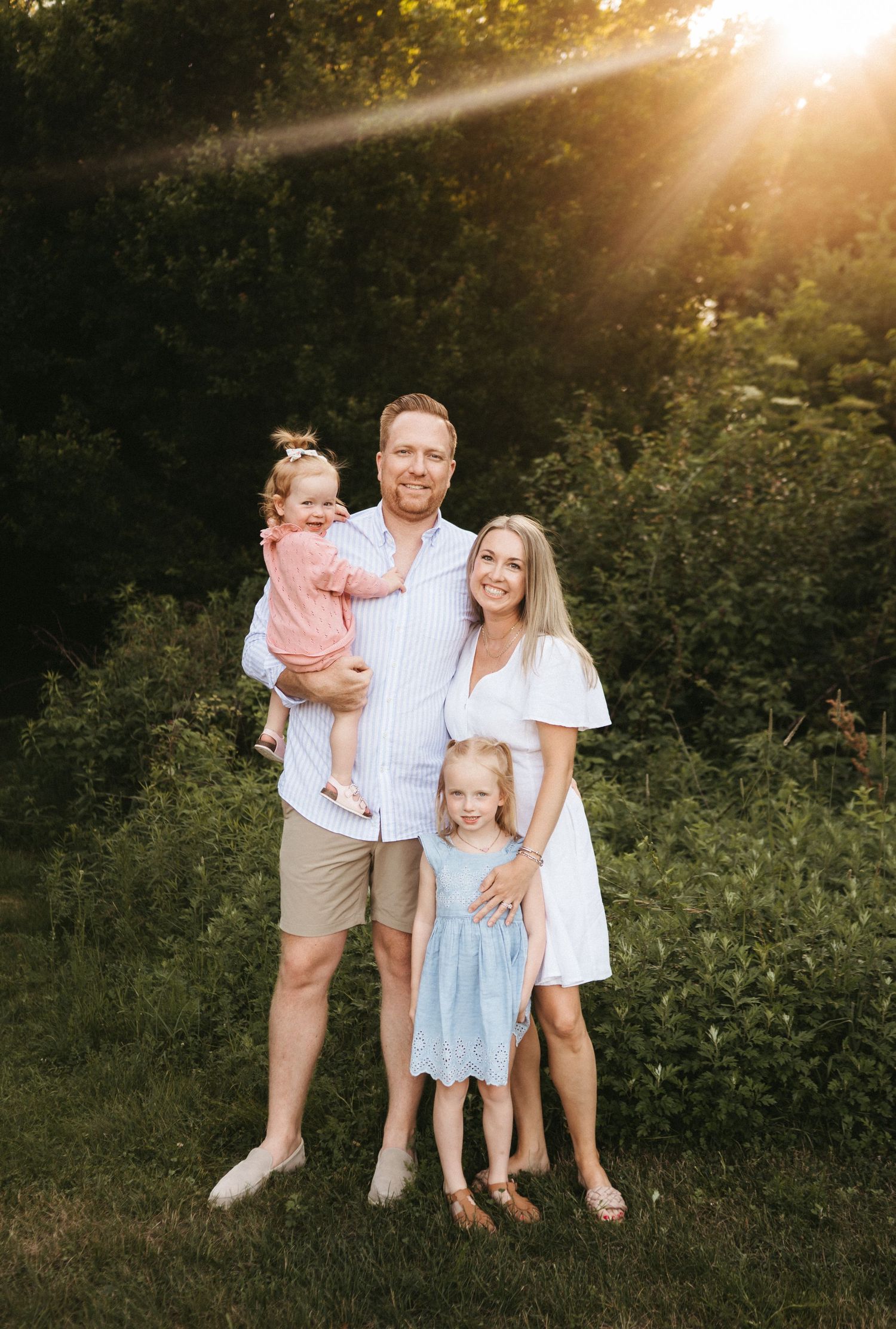 A family stands together in a natural outdoor setting with beautiful golden hour sunlight filtering through trees.