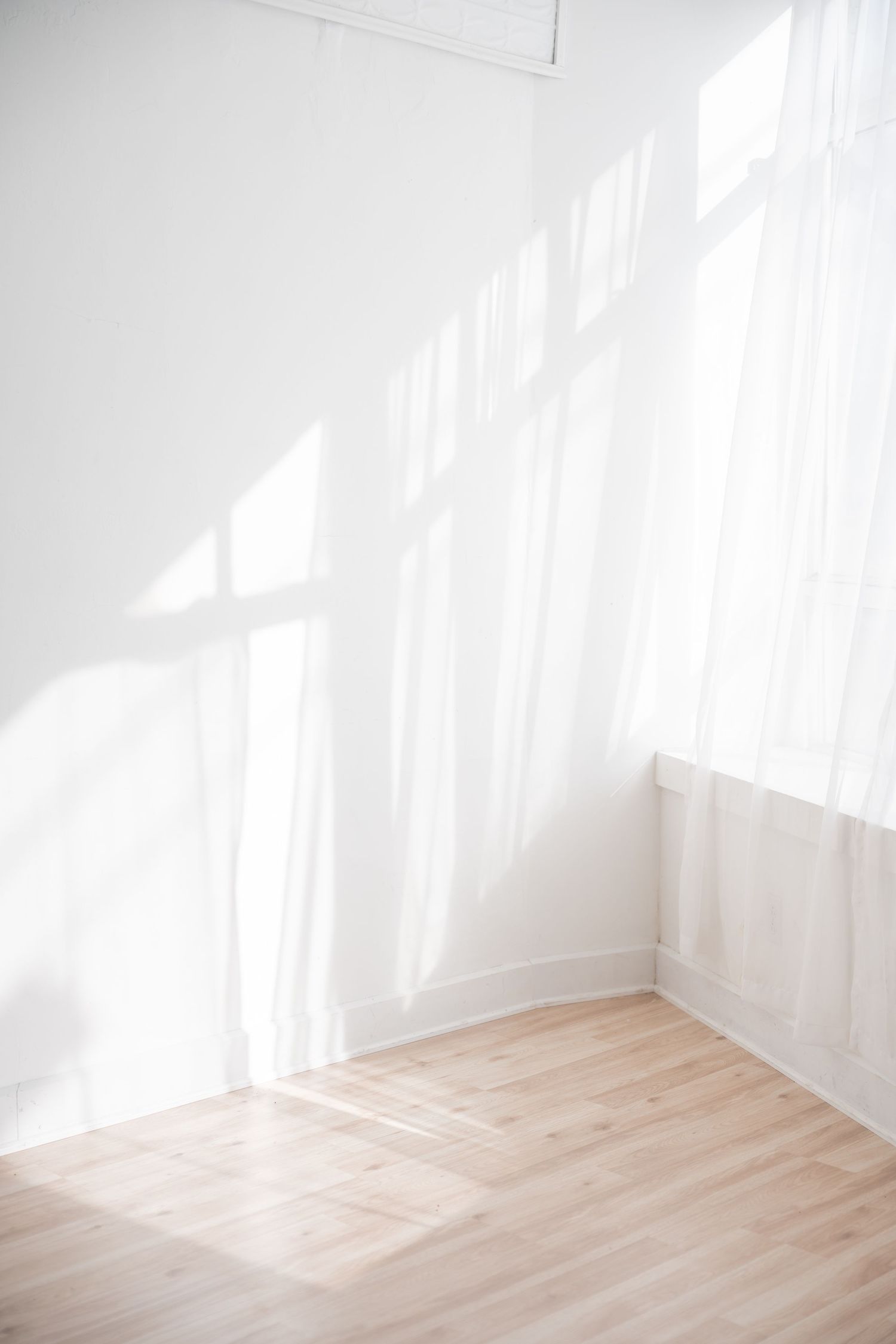 Bright sunlit room with white walls and hardwood flooring casting dramatic shadows through sheer curtains.