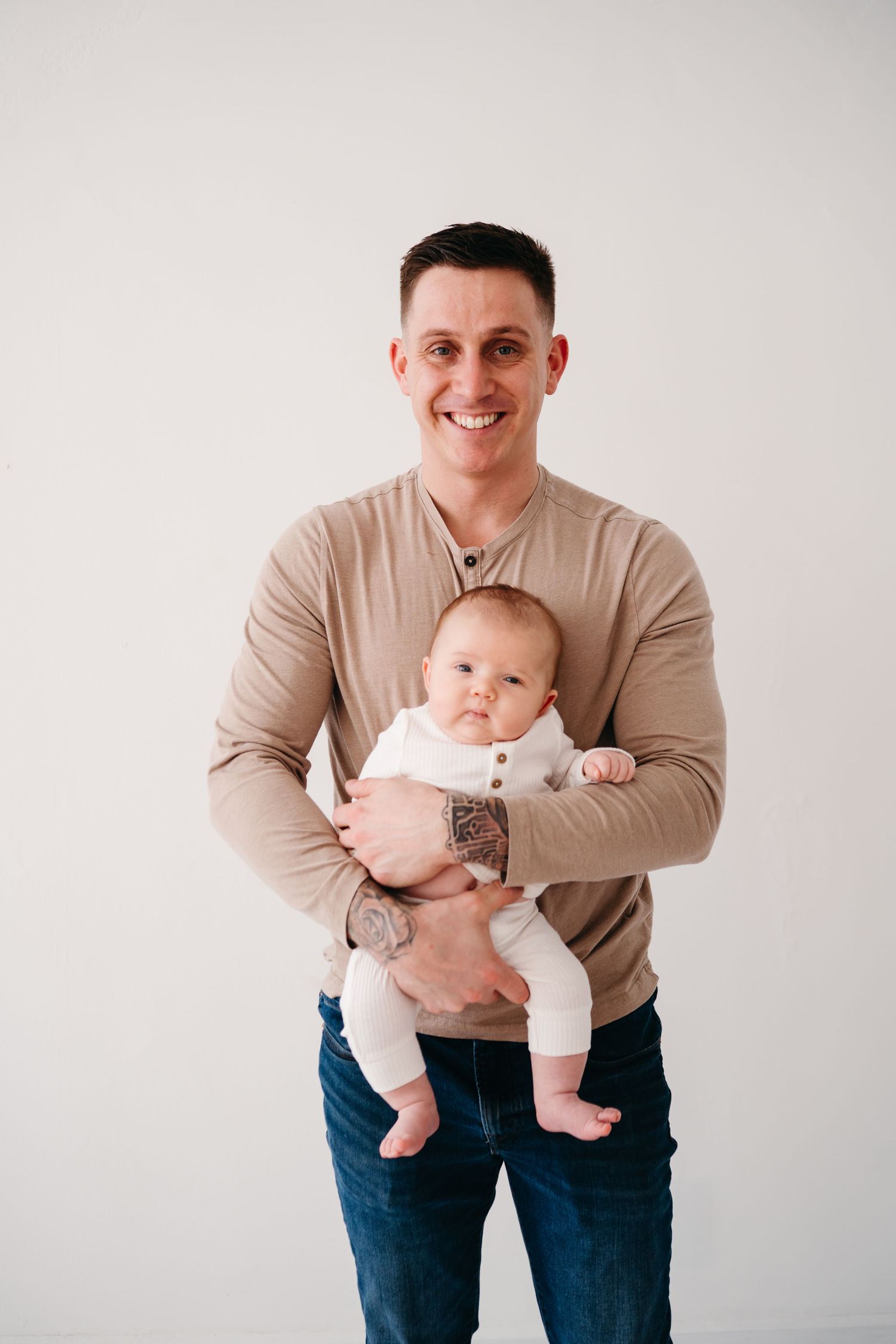 Adult in beige sweater poses with baby in white outfit against neutral background, both looking at camera.