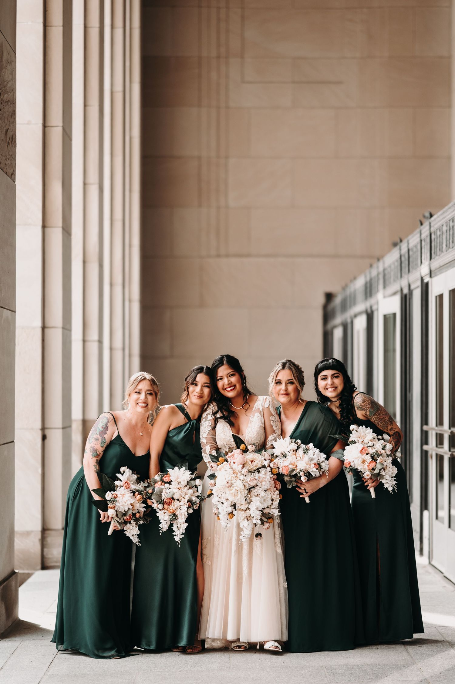 Bridesmaids in emerald green dresses pose with bouquets against classic stone architecture.