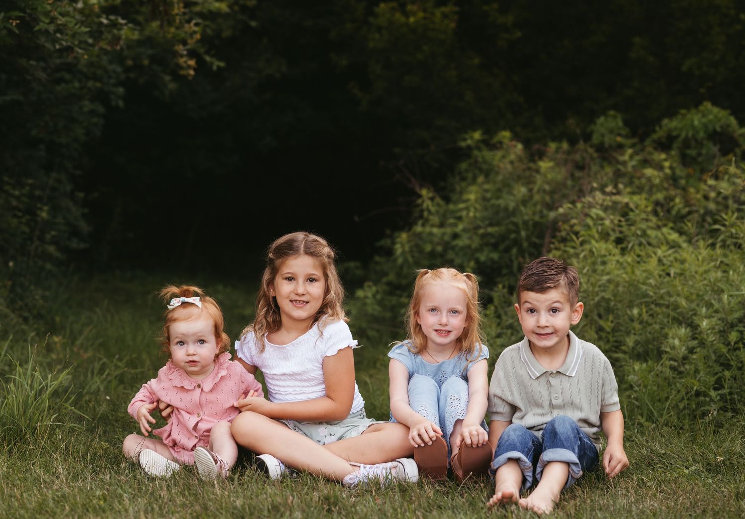 Four children sitting together on grass with natural greenery in the background.