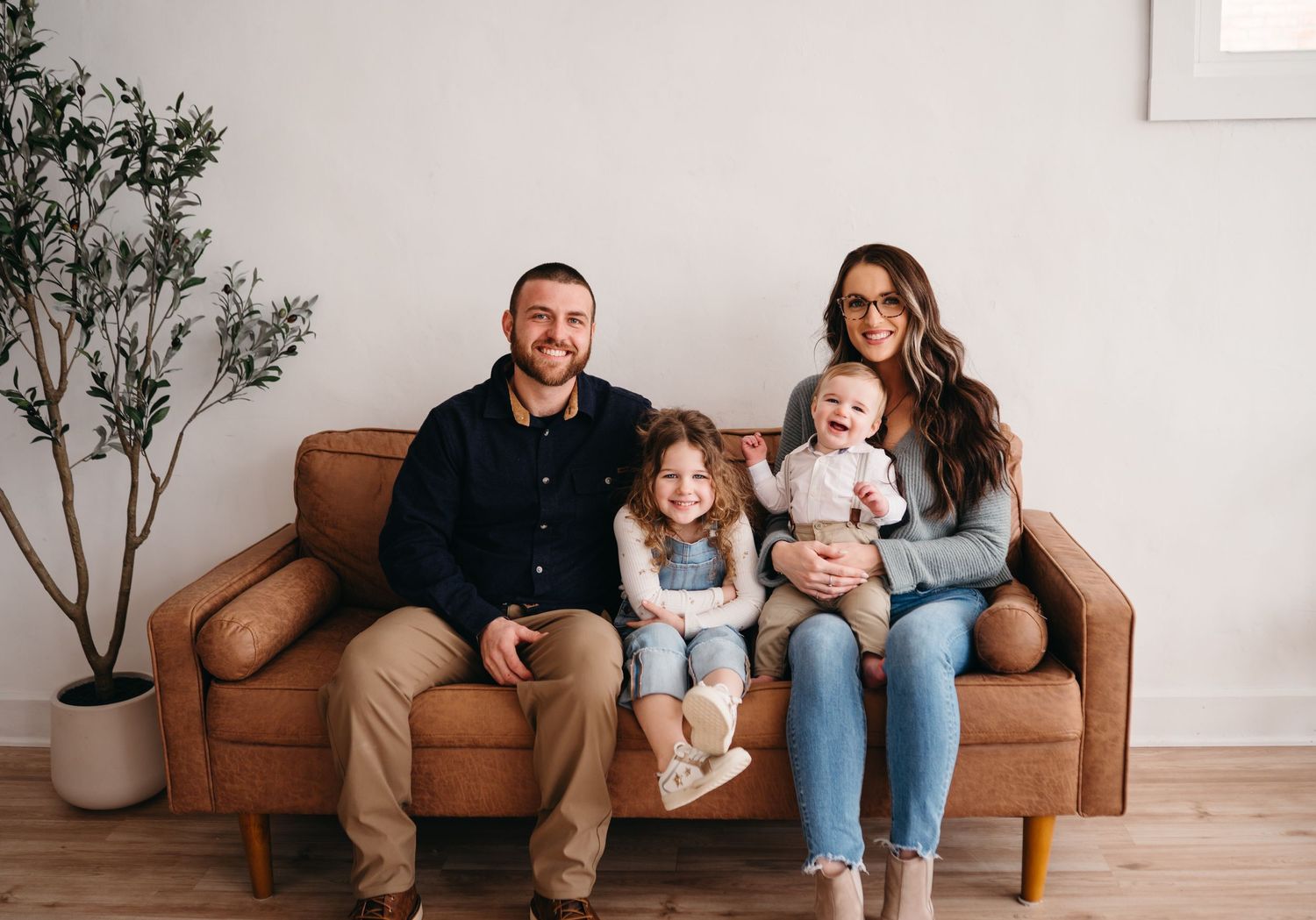 Family sits together on brown leather couch in minimalist living room with potted plant.