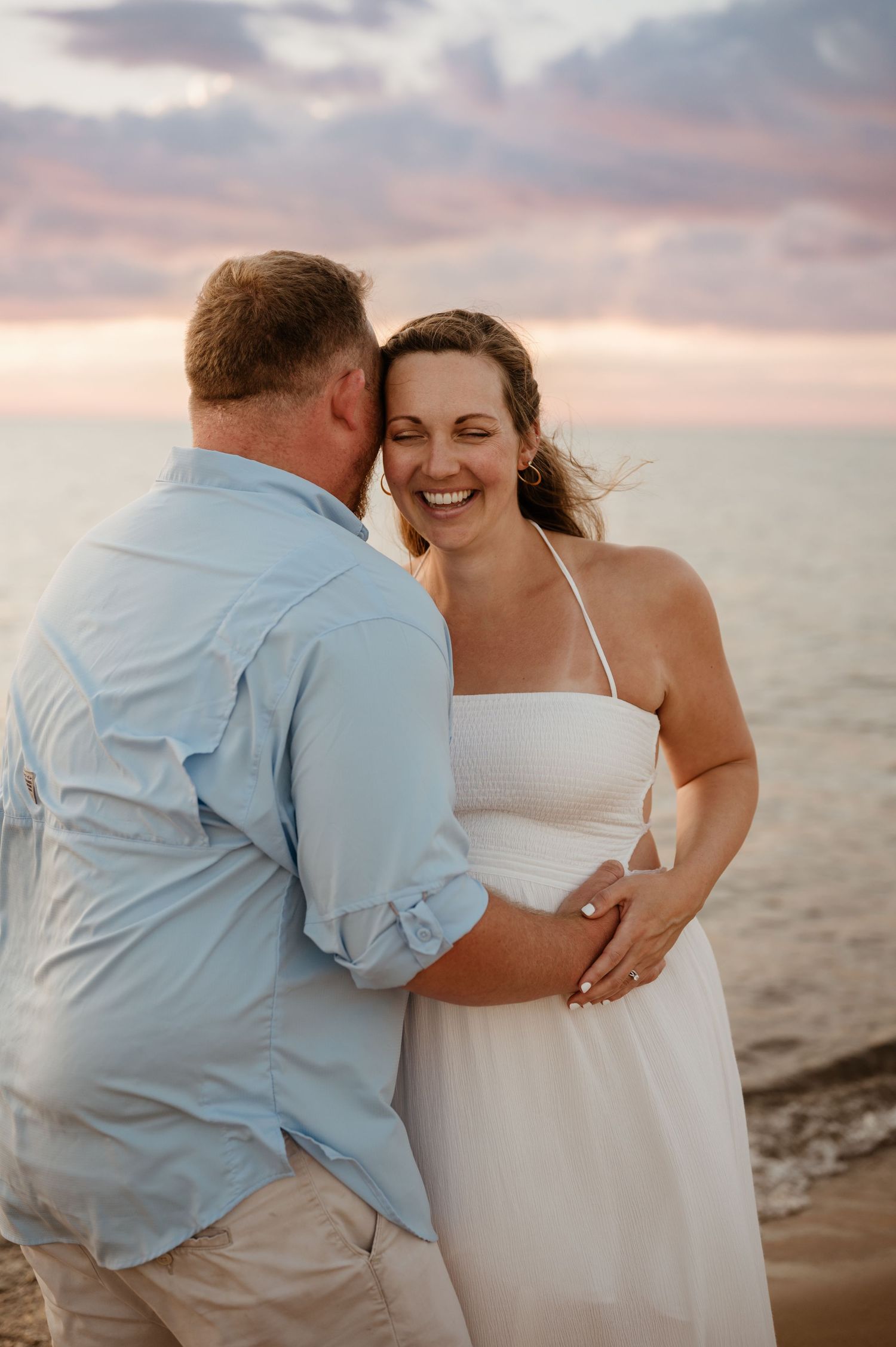A romantic beach scene at sunset with a couple embracing and laughing together on the sand.