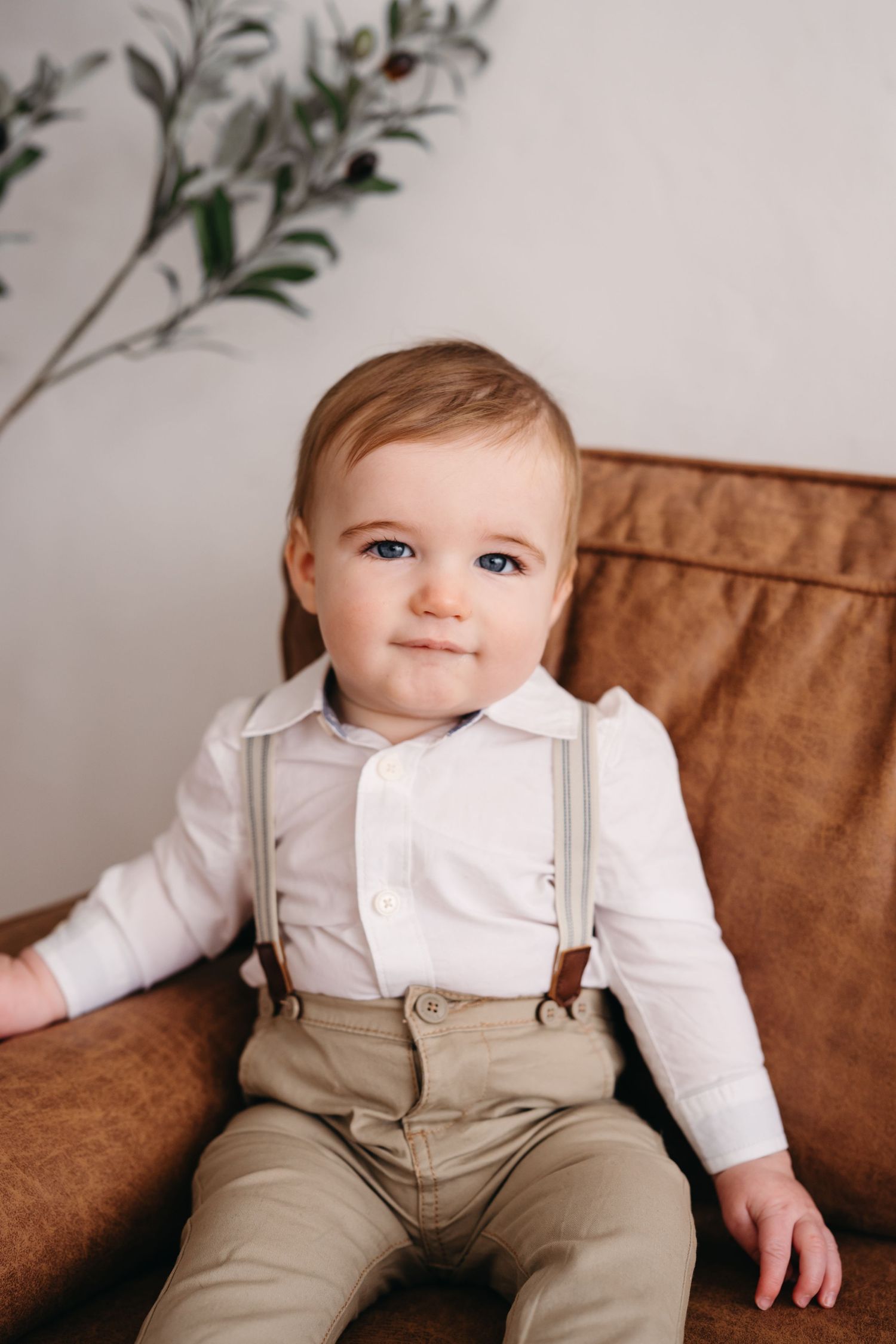 A baby in a white shirt and tan suspenders sits on a brown leather chair with greenery in the background.