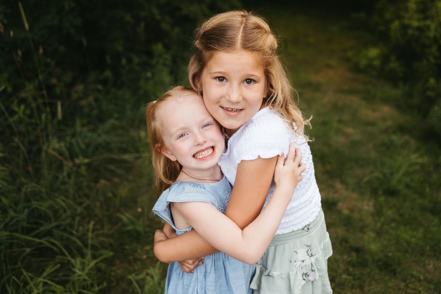 Two children share a joyful hug while standing in front of green foliage.