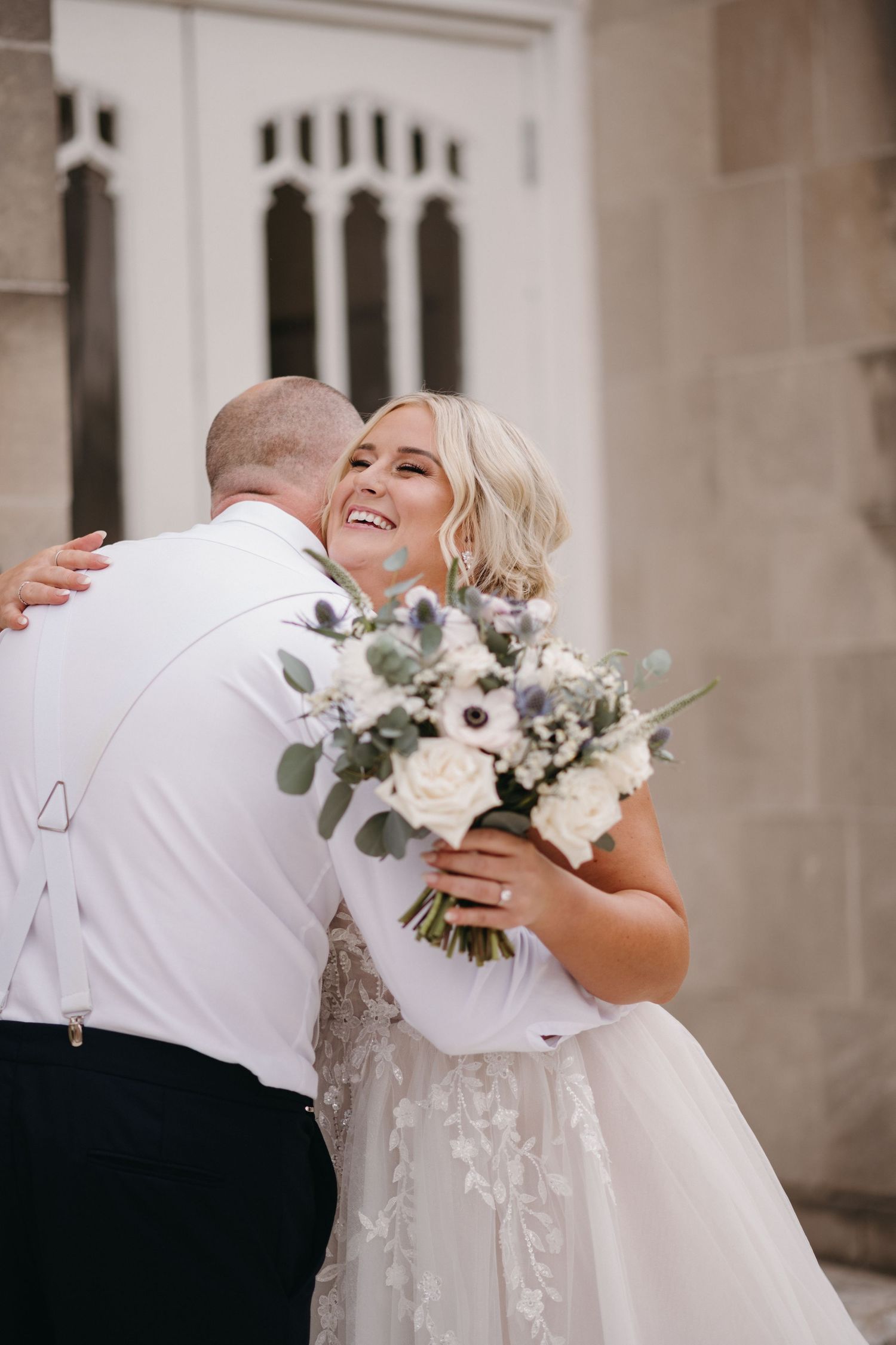 A bride in a lace gown embraces while holding a white and green bouquet outside a church.