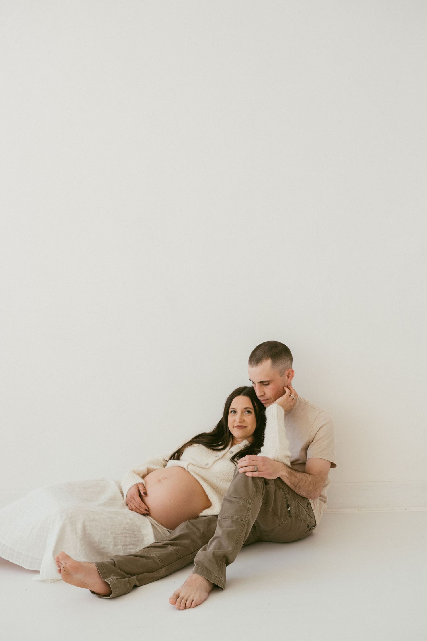 Couple in casual clothing sitting together against white wall during maternity photoshoot.