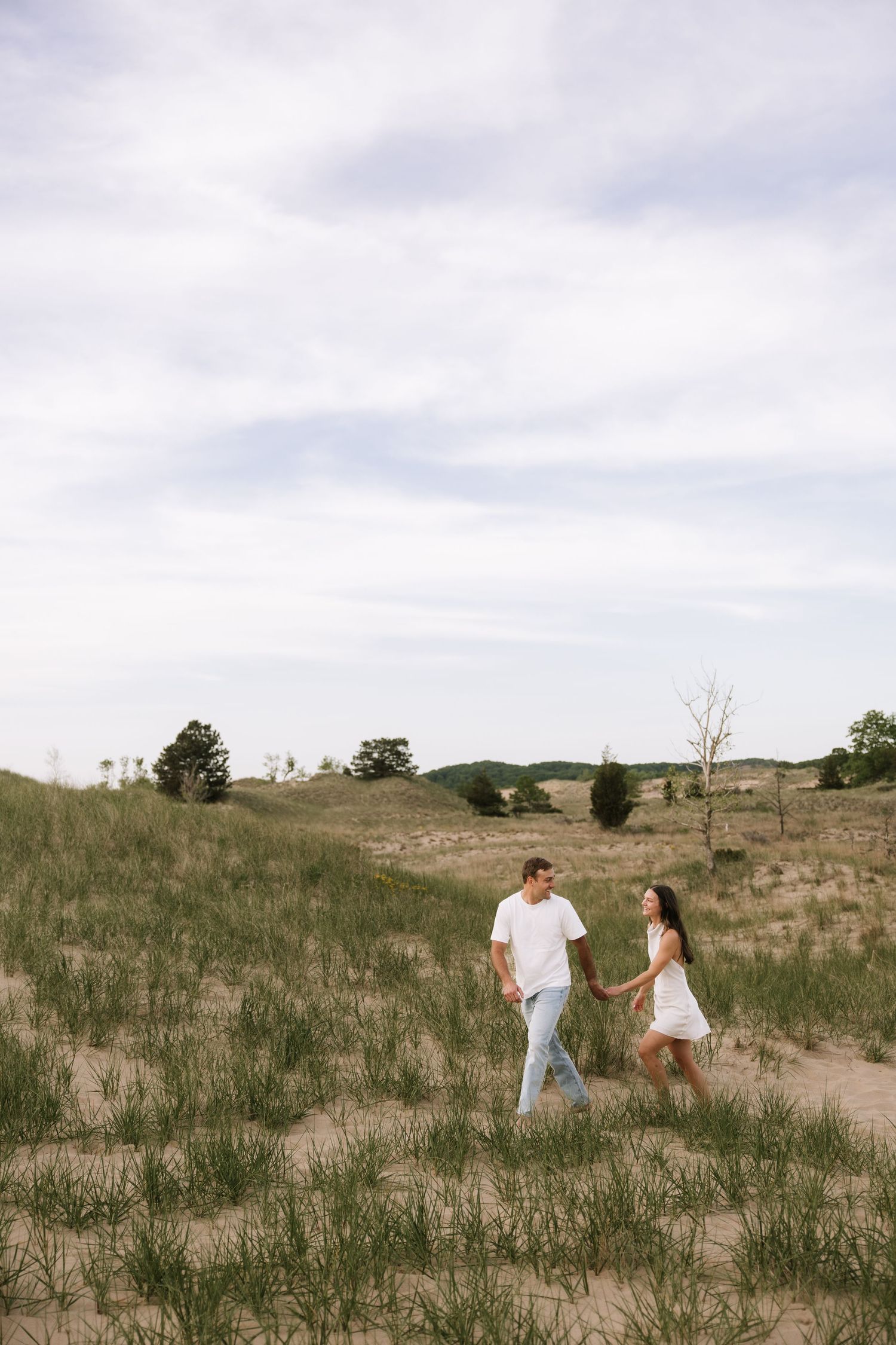 A couple in white outfits holds hands while walking through sandy dunes with green grass under a cloudy sky.