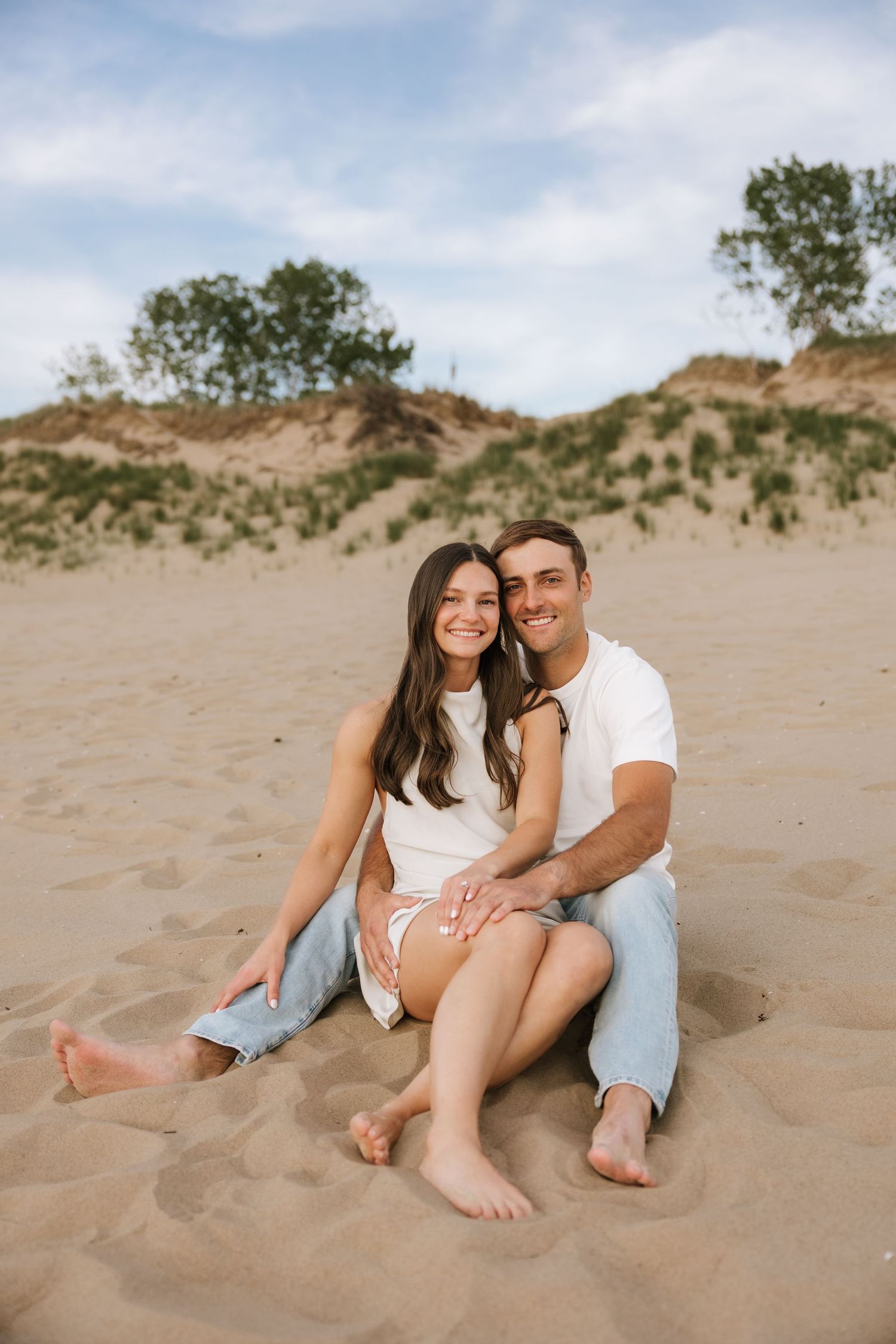 Smiling couple sitting together on sandy beach with sand dunes and green trees behind them.