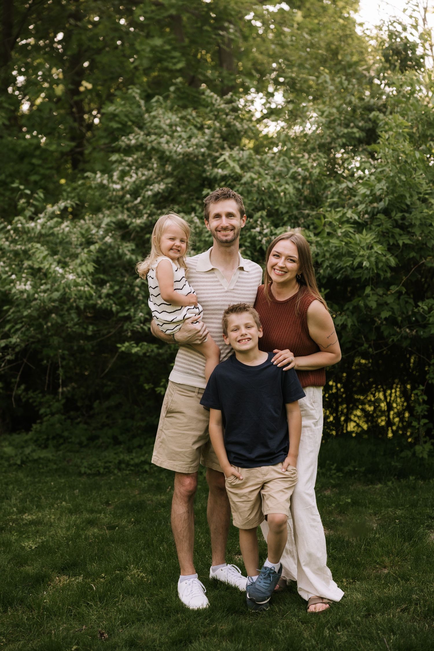 Happy family of four posing outdoors in lush green garden, parents with young son and toddler daughter.