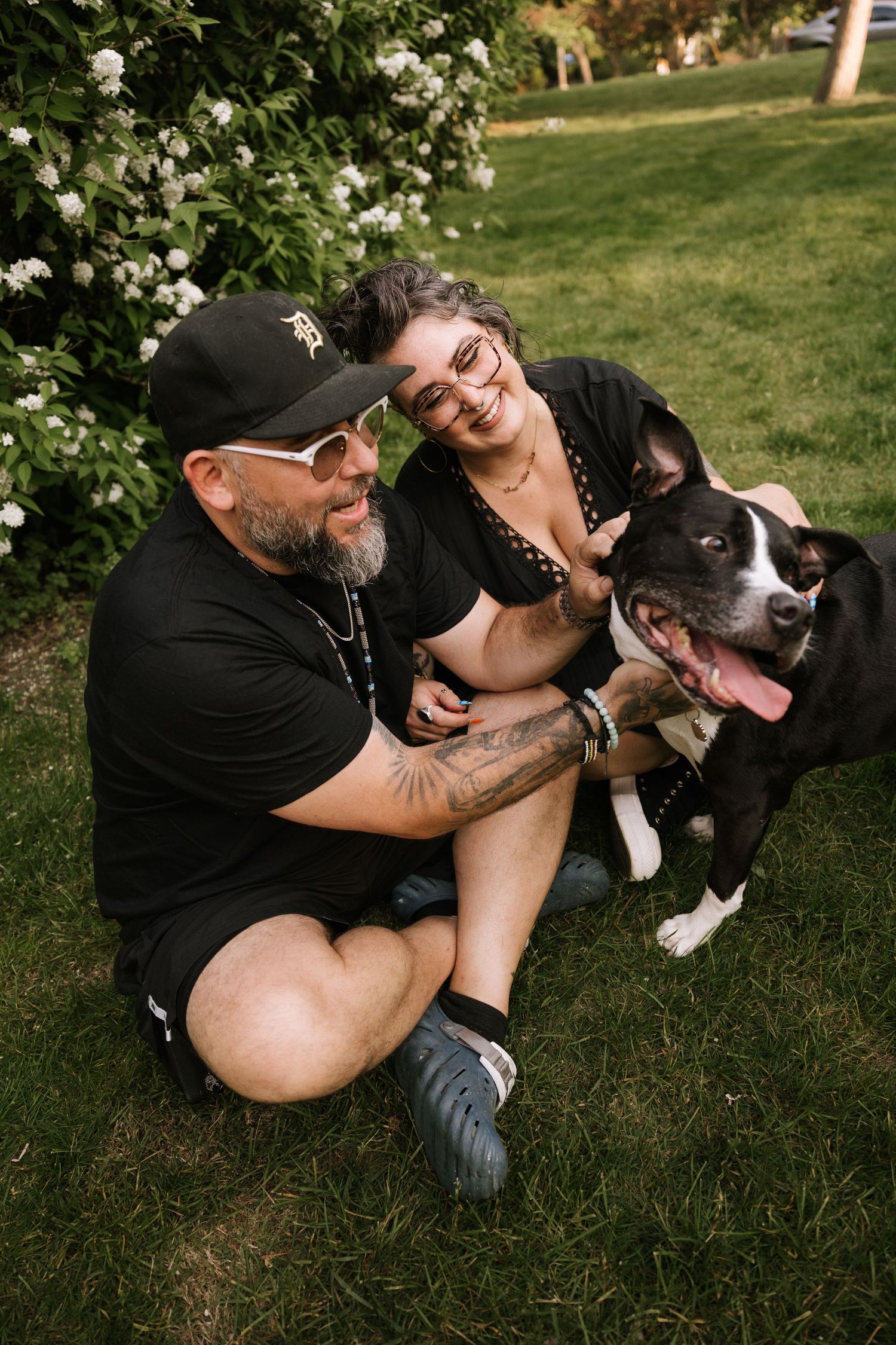 Couple sitting on grass with black and white dog near blooming white bushes outdoors.