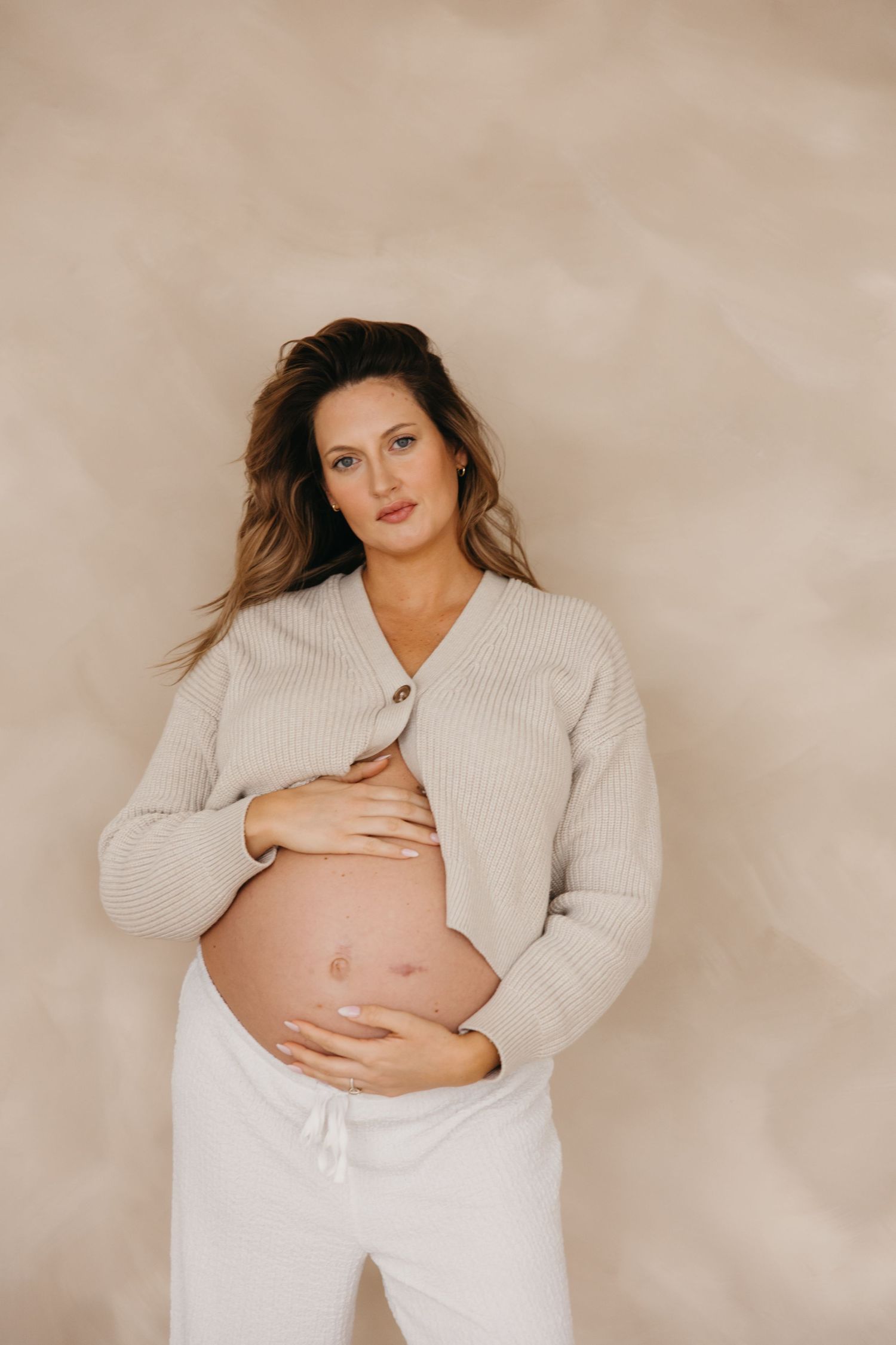 Pregnant woman in beige ribbed cardigan and white pants cradling bare baby bump against textured neutral backdrop.