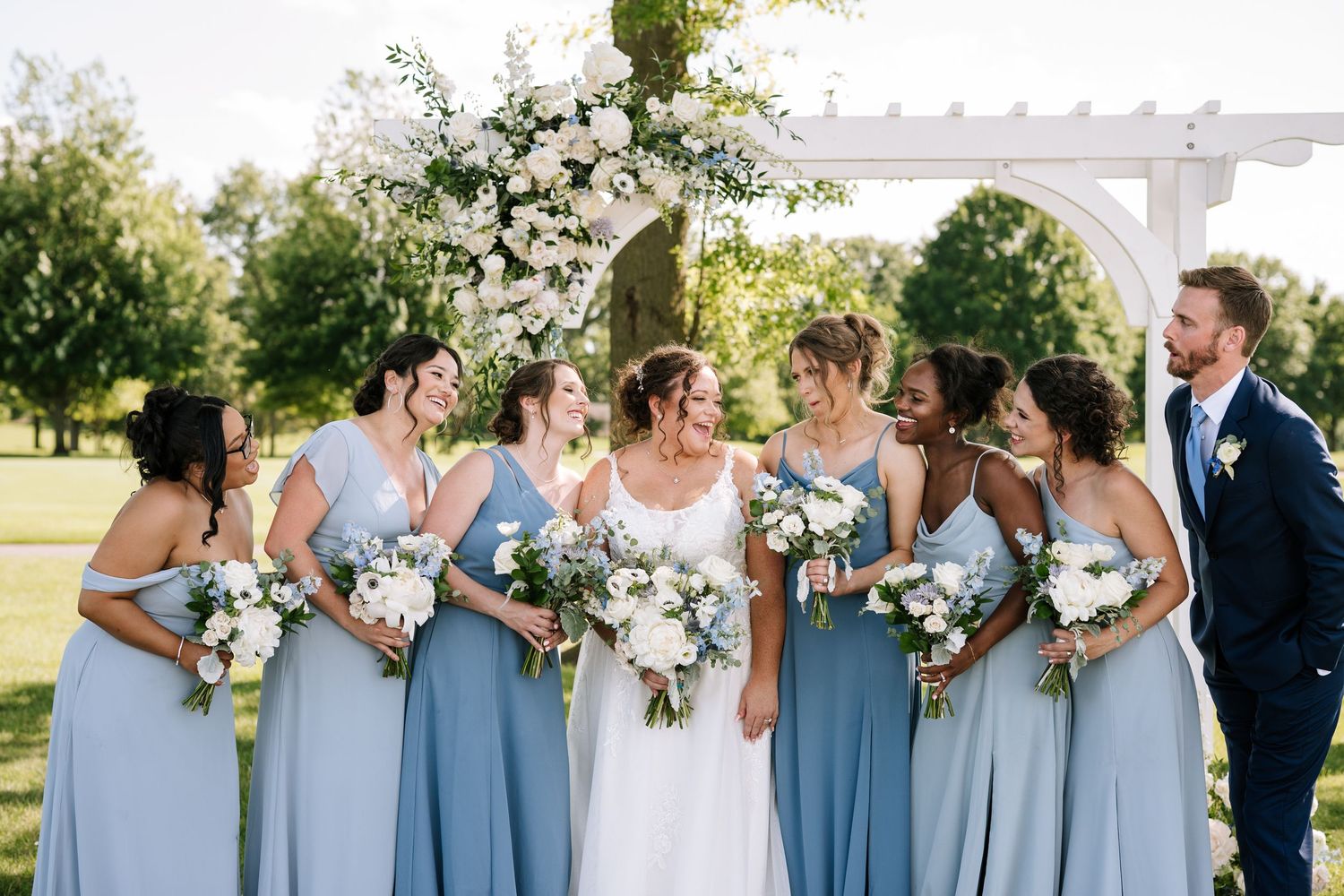 Wedding party in light blue bridesmaid dresses poses together under a white pergola.