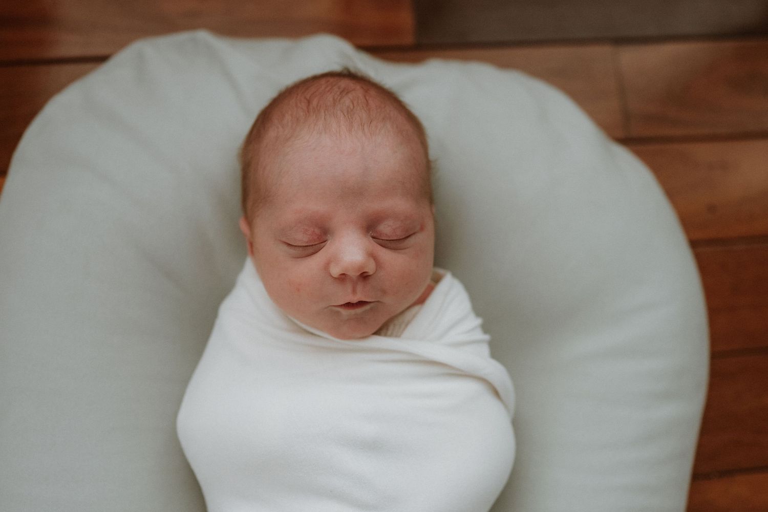 A sleeping newborn wrapped in white swaddle on a curved cushion against wooden floor.