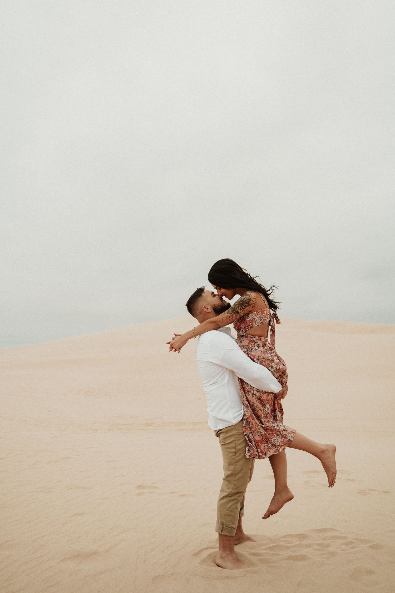 A romantic couple embraces and spins joyfully on sandy dunes against an overcast sky.
