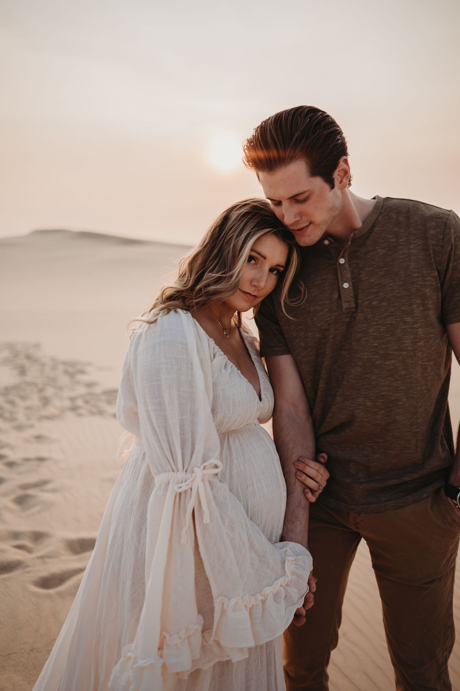 A tender moment between a couple in desert scenery with soft sunset lighting and rolling sand dunes.