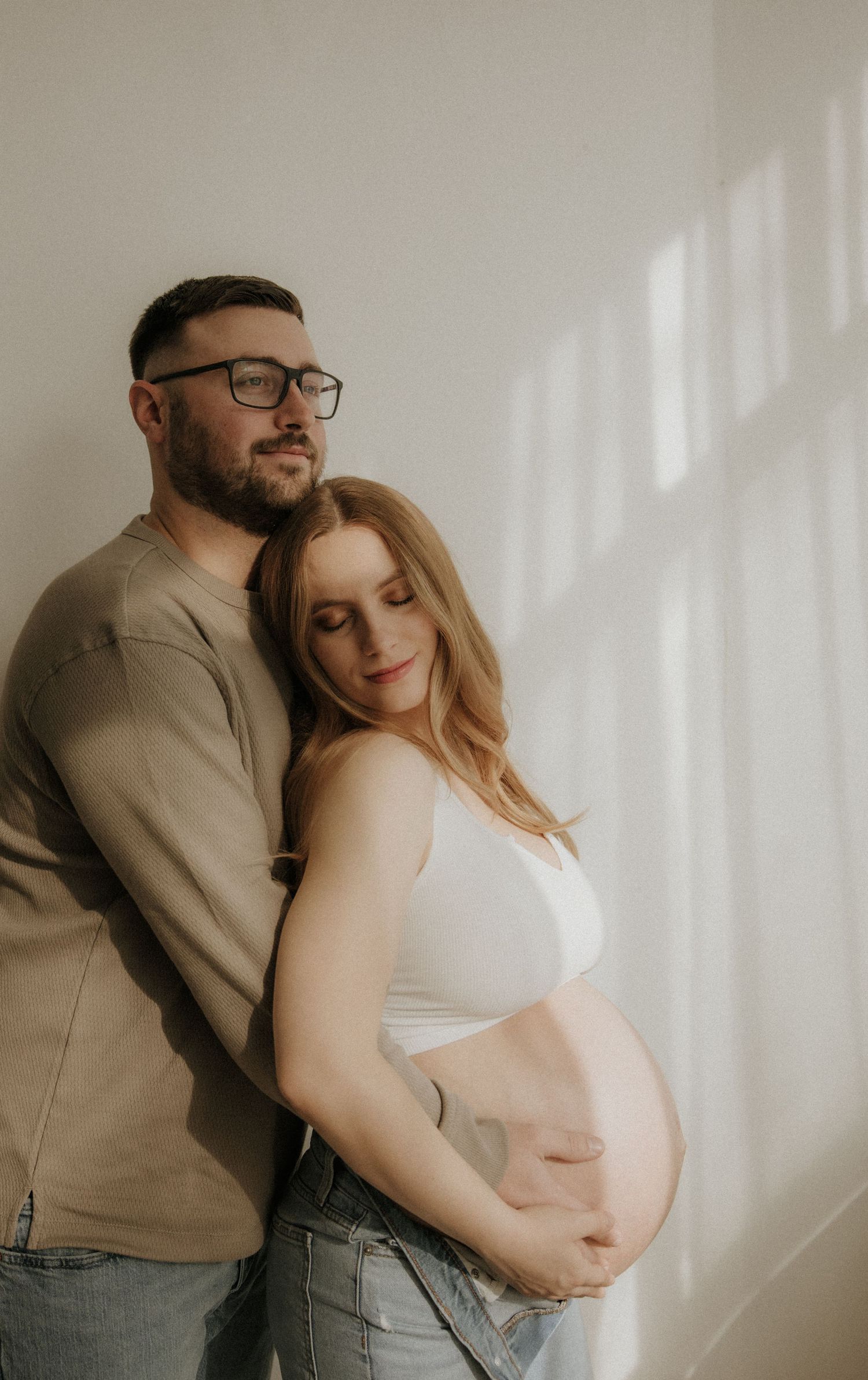 Intimate maternity portrait session with couple embracing against white wall.