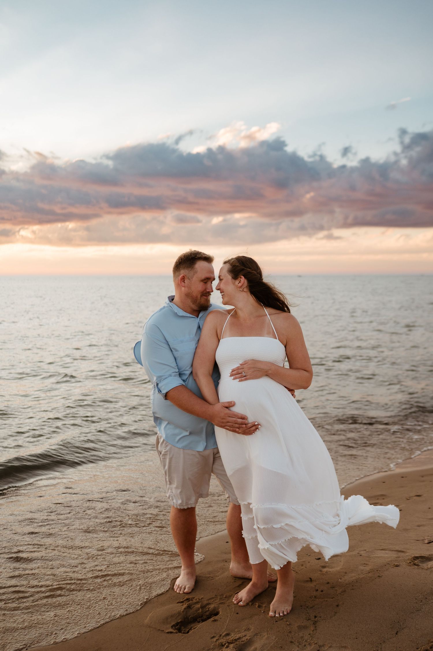 A romantic beach photoshoot at sunset with a couple embracing on the sand as waves roll in and clouds glow orange.