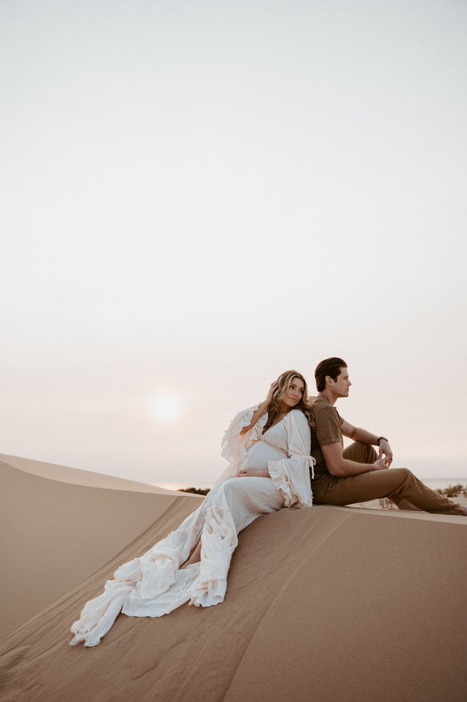 A couple in contrasting outfits sits back-to-back on a sand dune at sunset.
