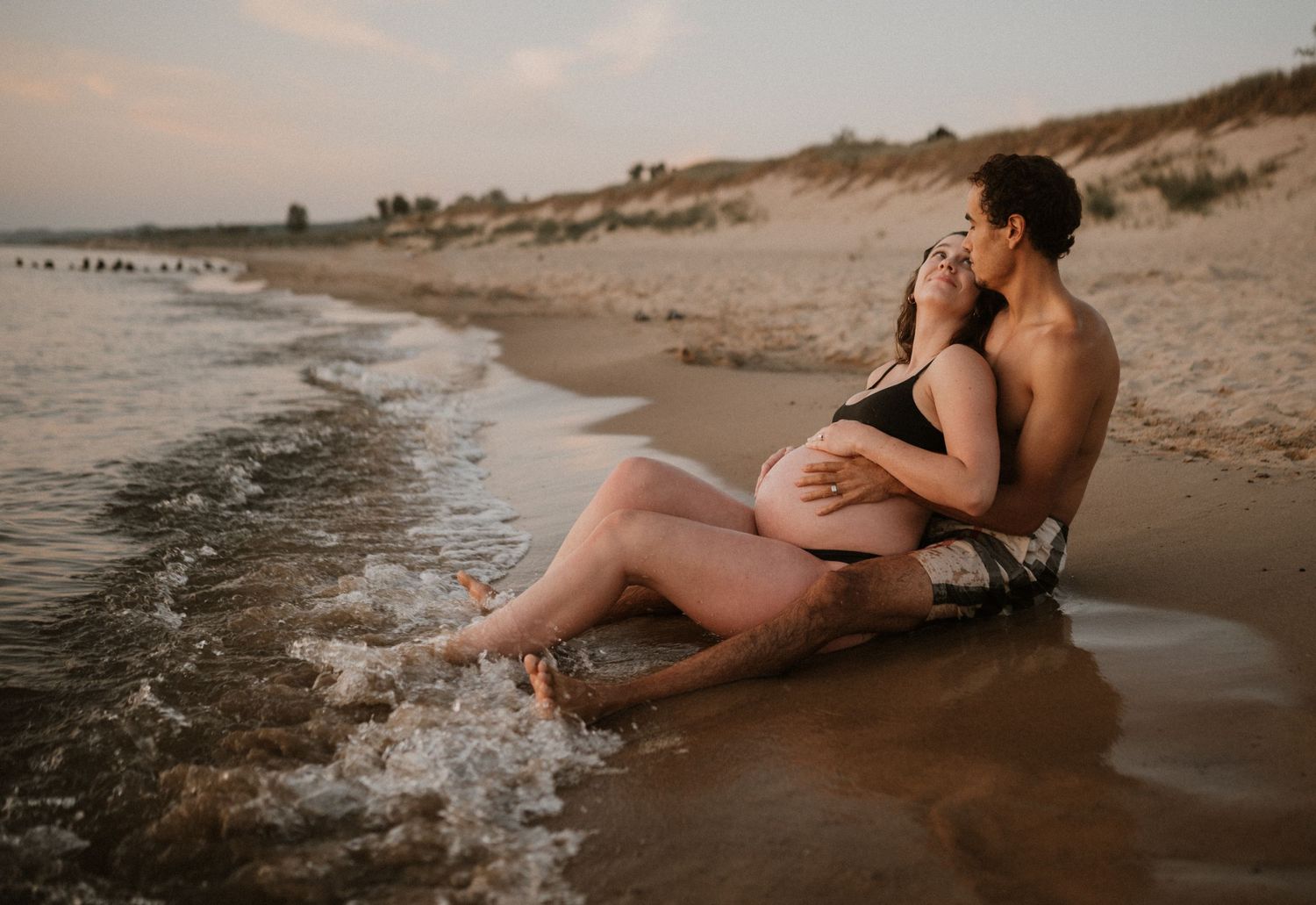 Couple embracing during sunset beach maternity session with waves lapping at their feet.