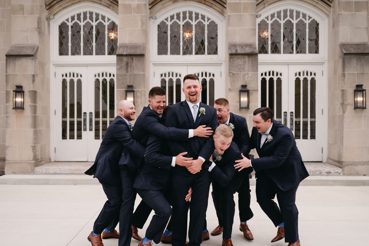 Groomsmen in dark suits playfully lift and carry one member of the group.