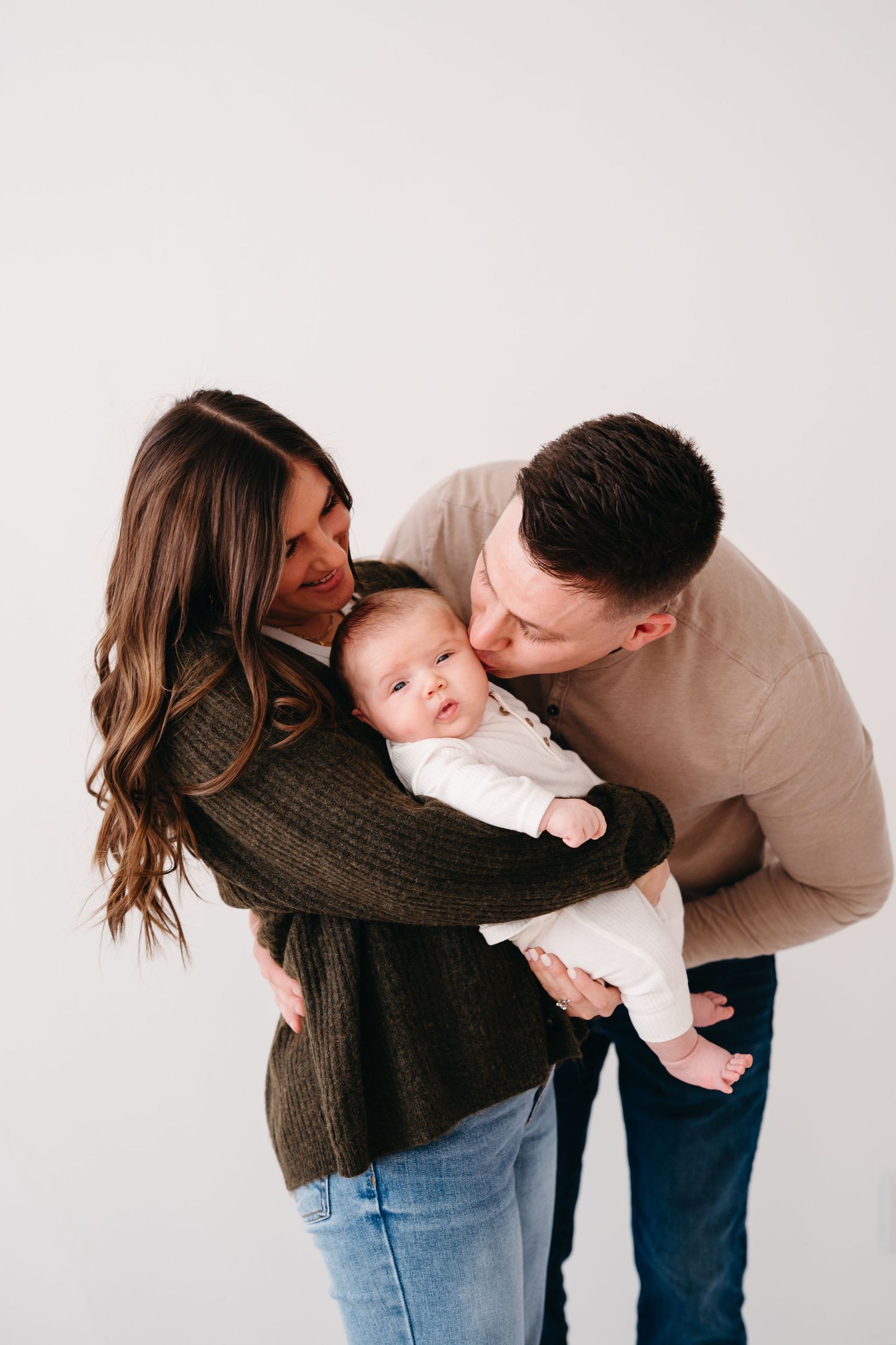 Parents share a tender moment kissing their baby while standing against a white background.