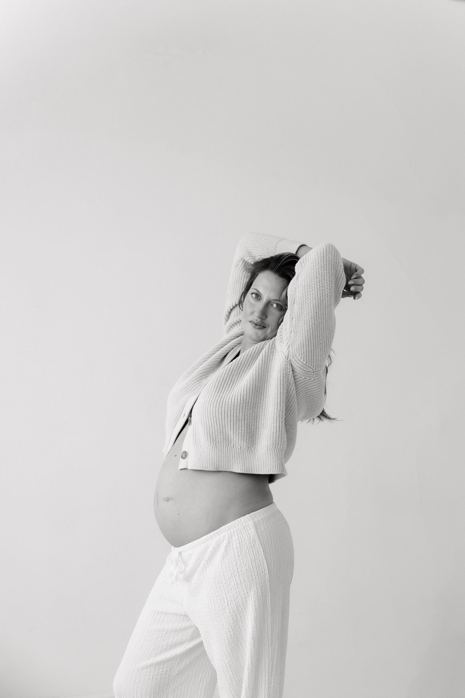 Black and white photo of pregnant woman in white outfit with arms raised, showing her baby bump.