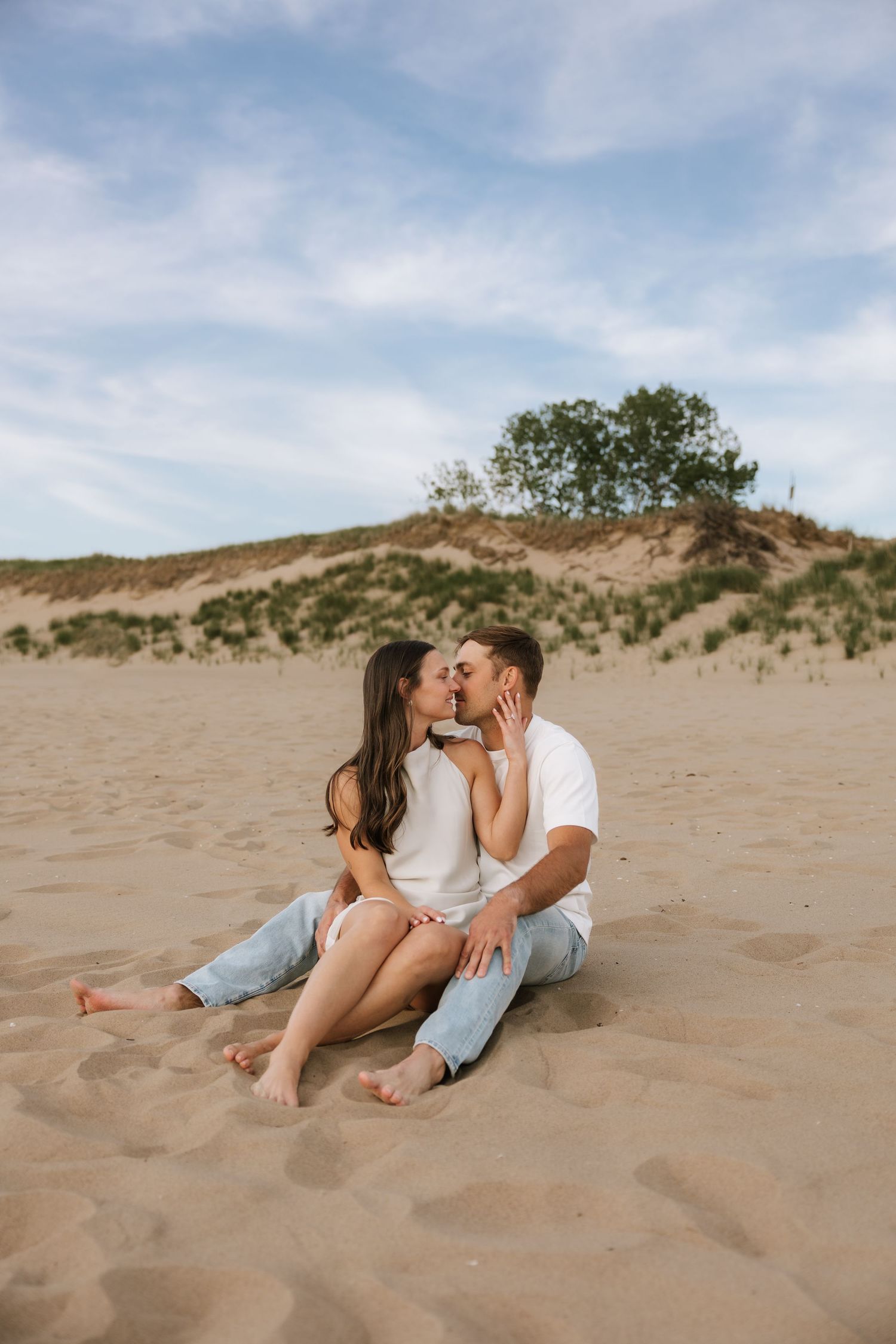 Couple sharing a romantic kiss while sitting together on sandy beach dunes with trees in the background.