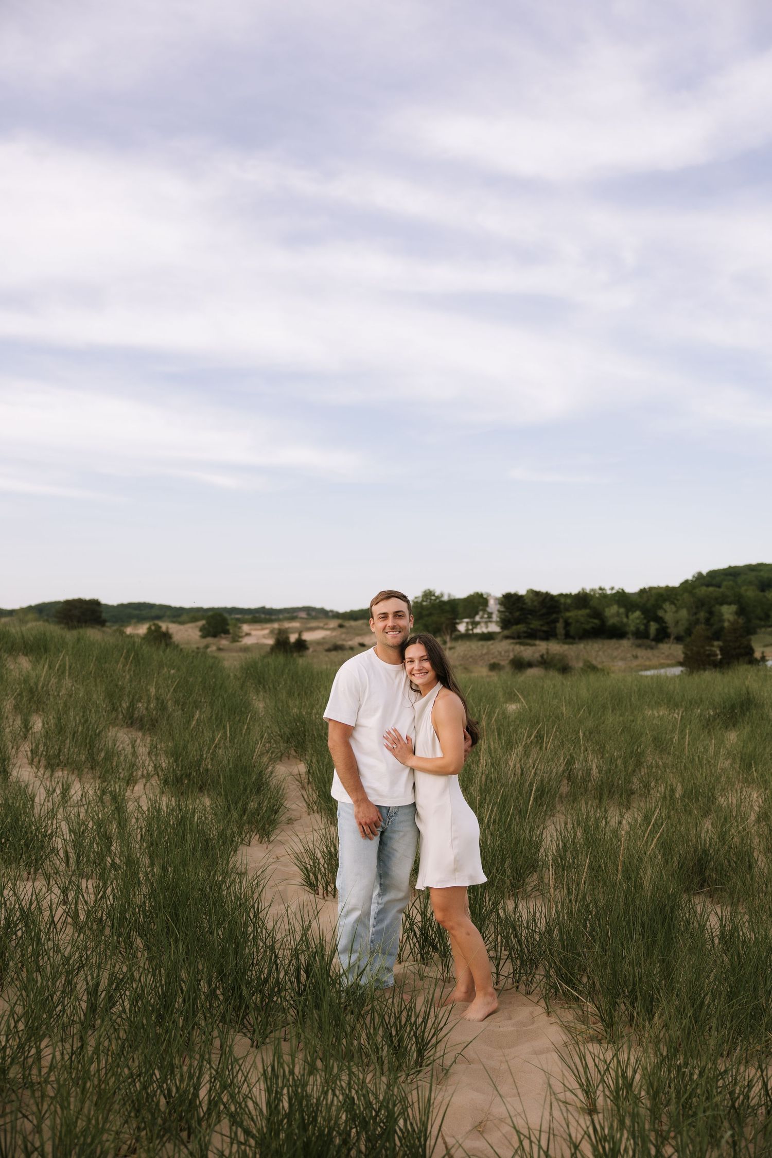 Happy couple embracing in sandy beach dunes with green grass, wearing white outfits on a cloudy day.