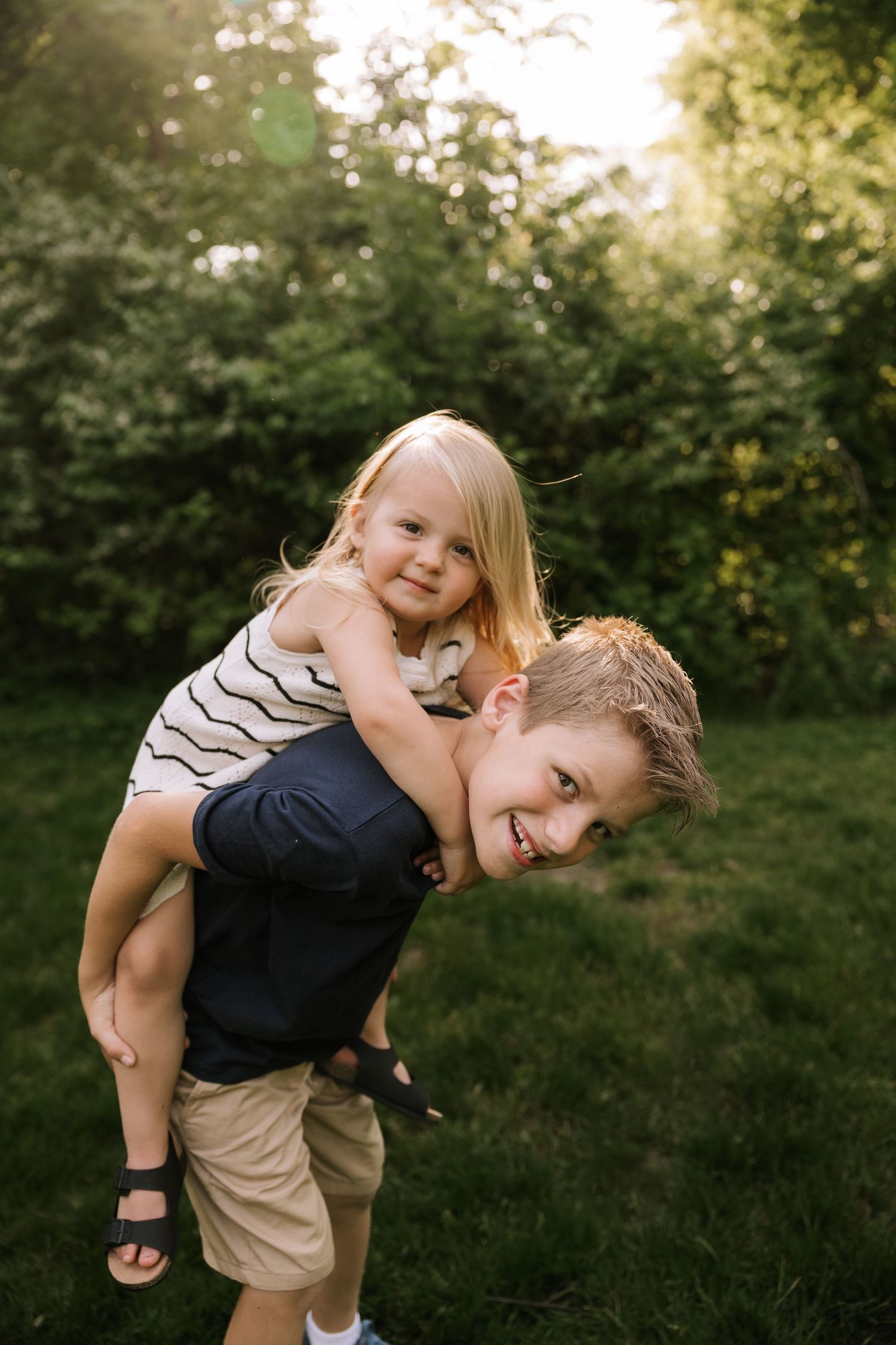A smiling boy gives his little blonde sister a piggyback ride in a sunny green backyard.