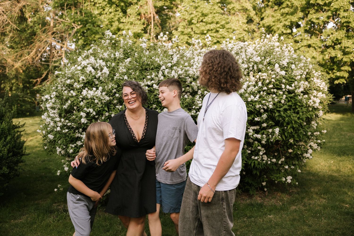 A joyful family of four laughs together outdoors near blooming white bushes in a lush green garden.