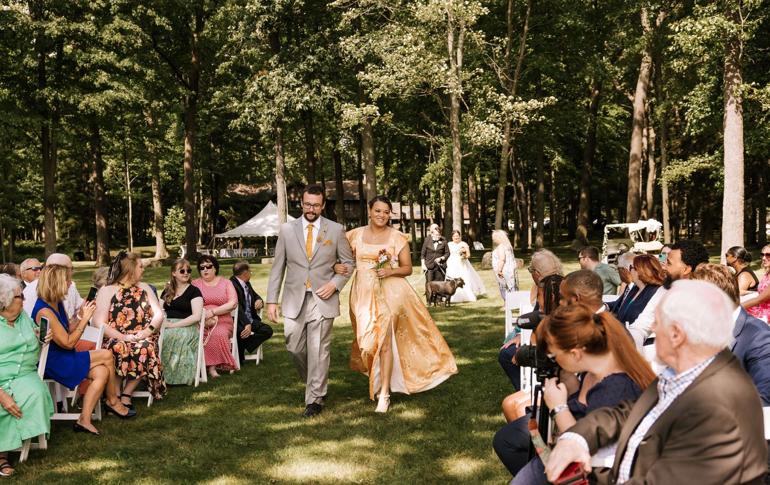 Wedding guests gather outdoors in a wooded setting for a celebration on a sunny summer day.