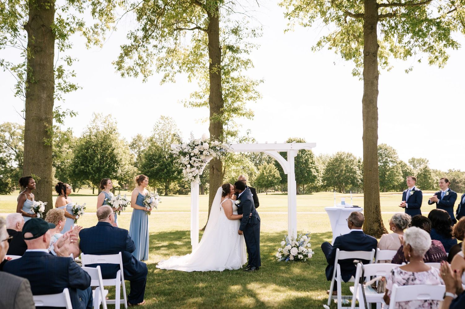 Outdoor wedding ceremony under tall trees with white chairs and floral arch.