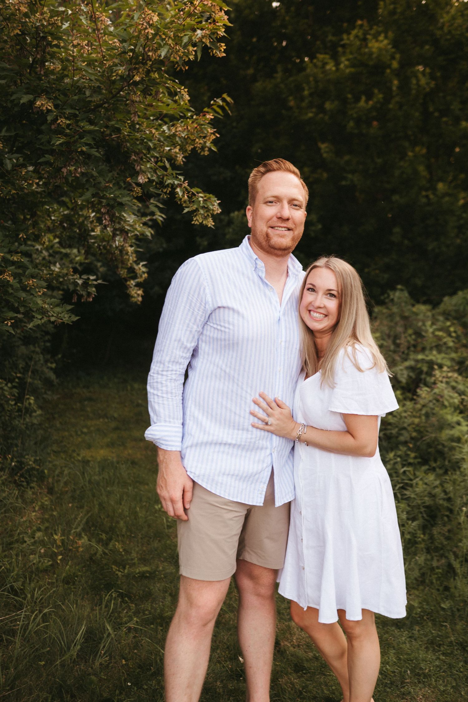 A couple poses outdoors in coordinated white and beige summer outfits against a lush green backdrop.