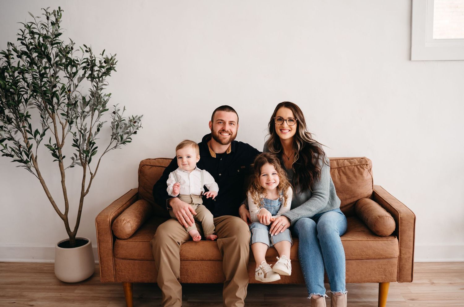 A family relaxing together on a tan leather couch in a minimalist living room with a potted plant.