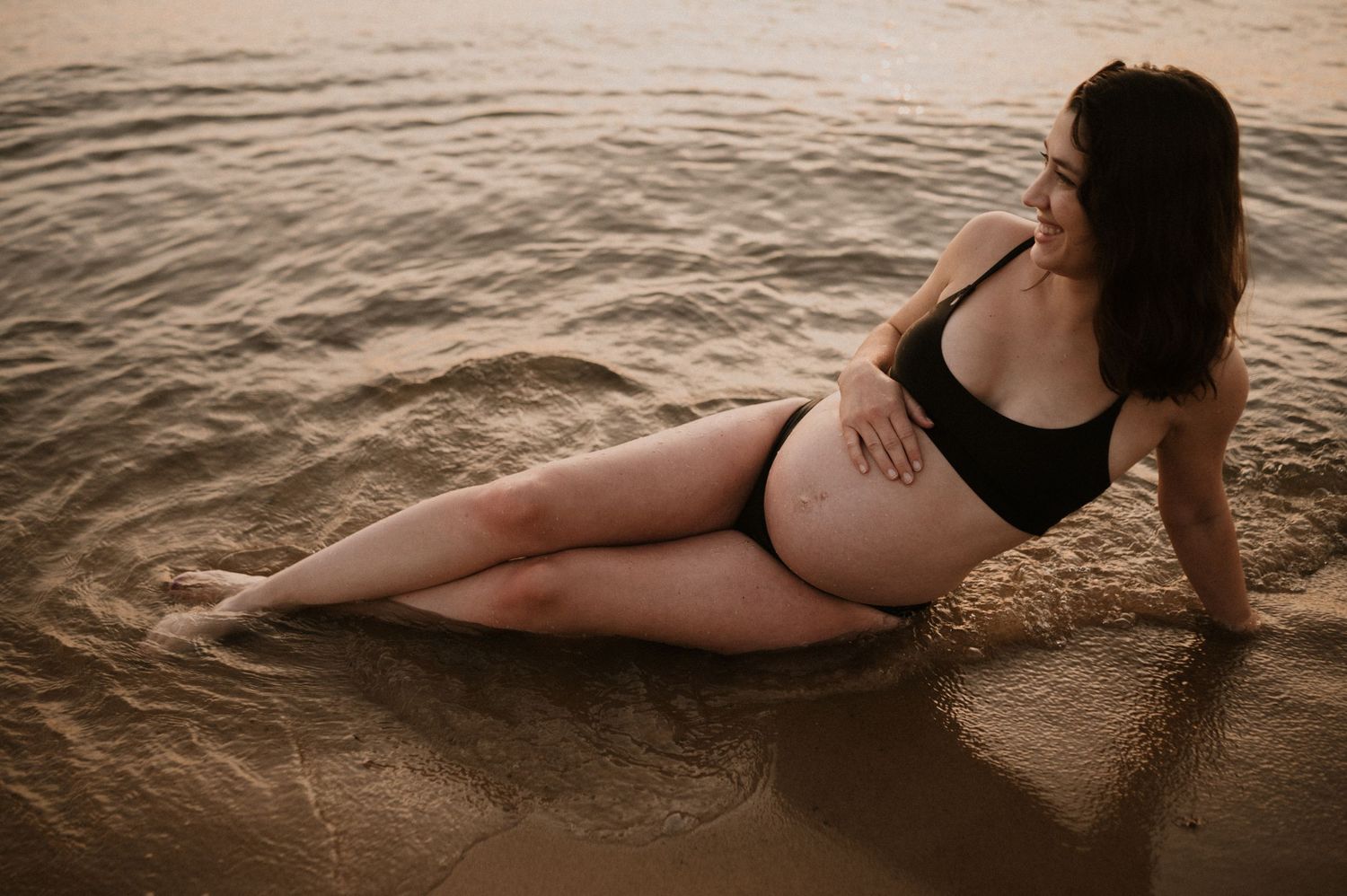 Maternity beach photo of woman in black swimwear posing at water's edge.