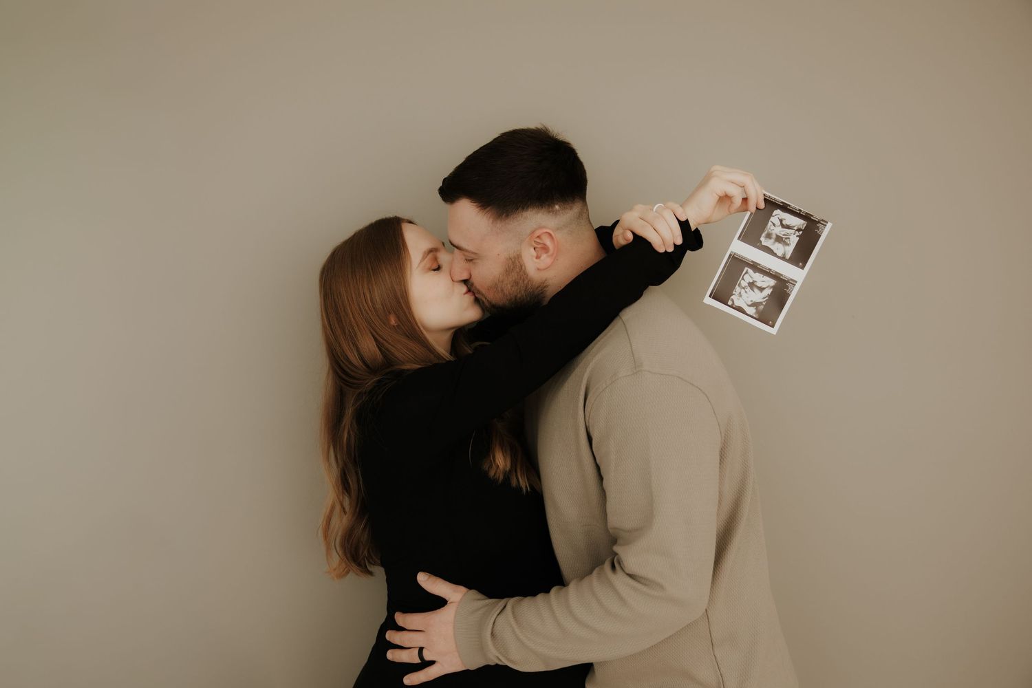 A couple embraces while holding ultrasound photos against a neutral wall background.