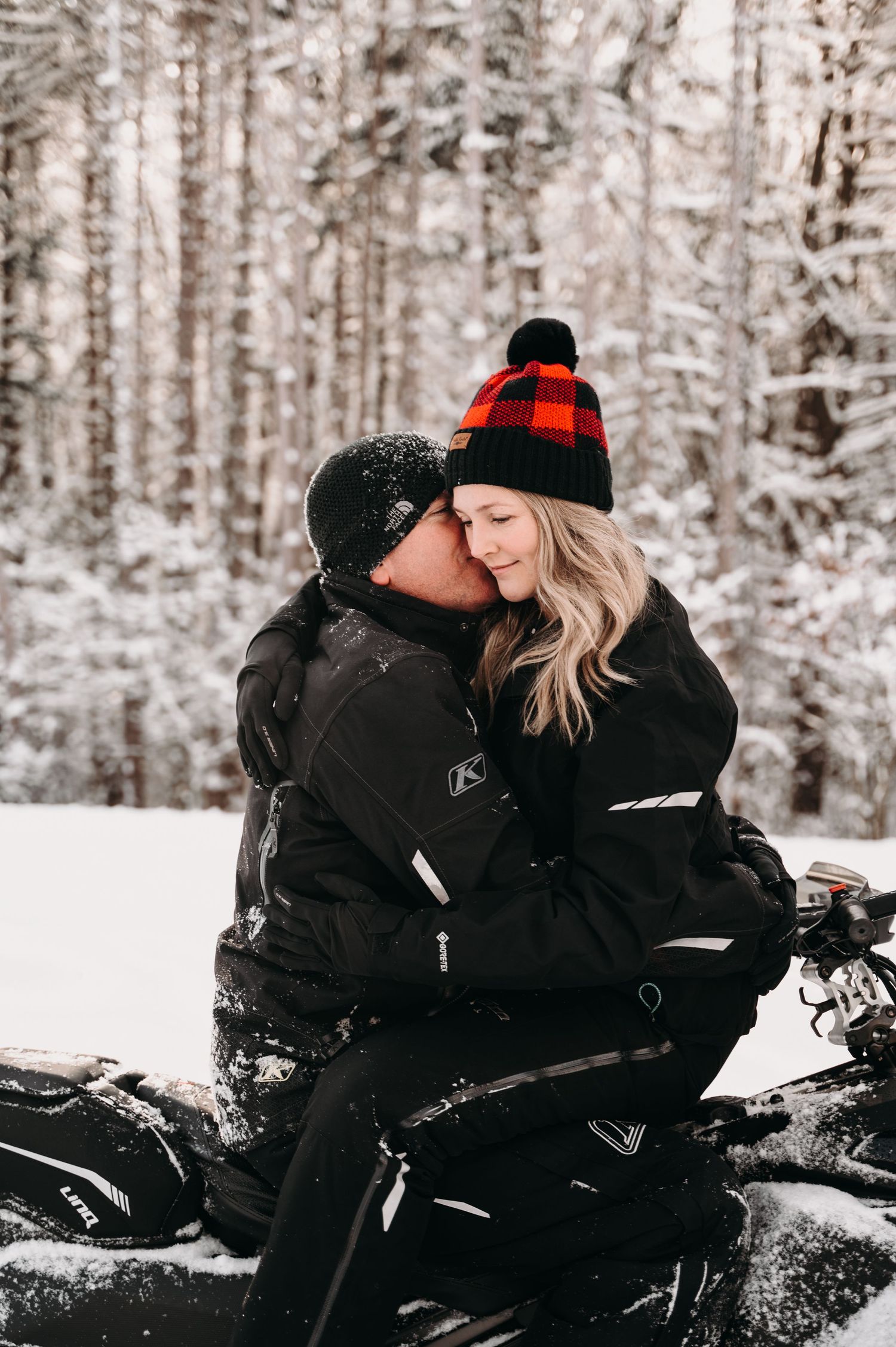 Couple in winter gear sharing an intimate moment on a snowmobile in a snowy forest.